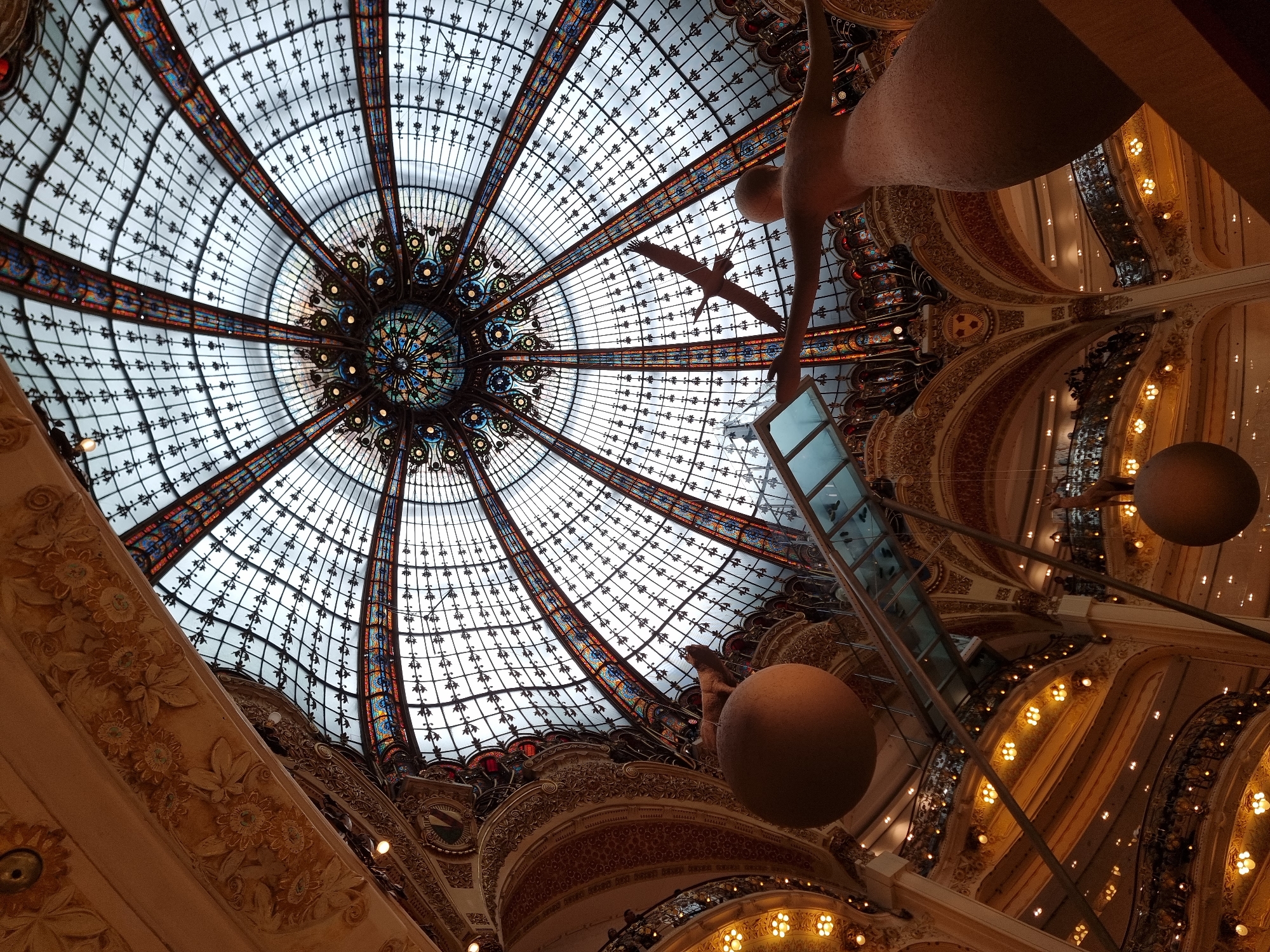 The image depicts the interior of Galeries Lafayette Haussmann in Paris, France, specifically the stained-glass dome. The dome is intricate with geometric patterns and vibrant colors. Underneath the dome, architectural details of the building are visible, including ornate balconies and a partially visible sculpture of a woman. Spherical ornaments and a glass-encased elevator or installation are suspended in the space. The angle of the photograph is looking upwards.