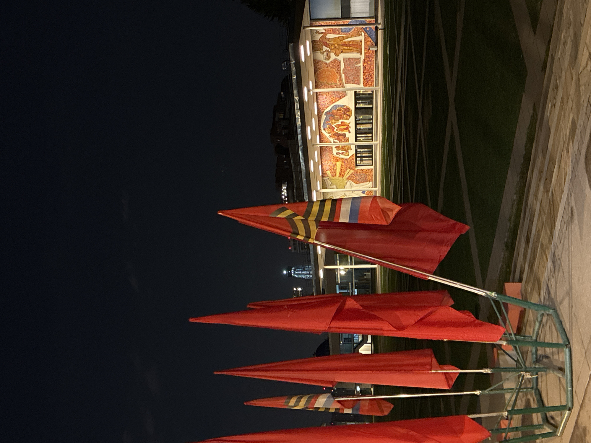 A low-angle, medium-shot photograph shows a set of red pennant flags, along with what appears to be a mural on the side of a building in the city of Unknown, Unknown.
The photograph is taken at night, with the dark sky providing a stark backdrop. In the foreground, a series of red pennant flags, arranged in a multi-tiered display, dominates the view. The flags, positioned vertically, are triangular in shape and appear to be made of a synthetic material. Some of the flags have stripe details at the top. The flags are attached to a green metal frame.
Behind the flags, the mural on the side of the building is illuminated, revealing an abstract design. The mural appears to be made of tiles. The building with the mural has multiple stories and windows.
The lighting in the scene suggests artificial light sources illuminating the flags and mural. The environment includes concrete pavement, along with grass areas.