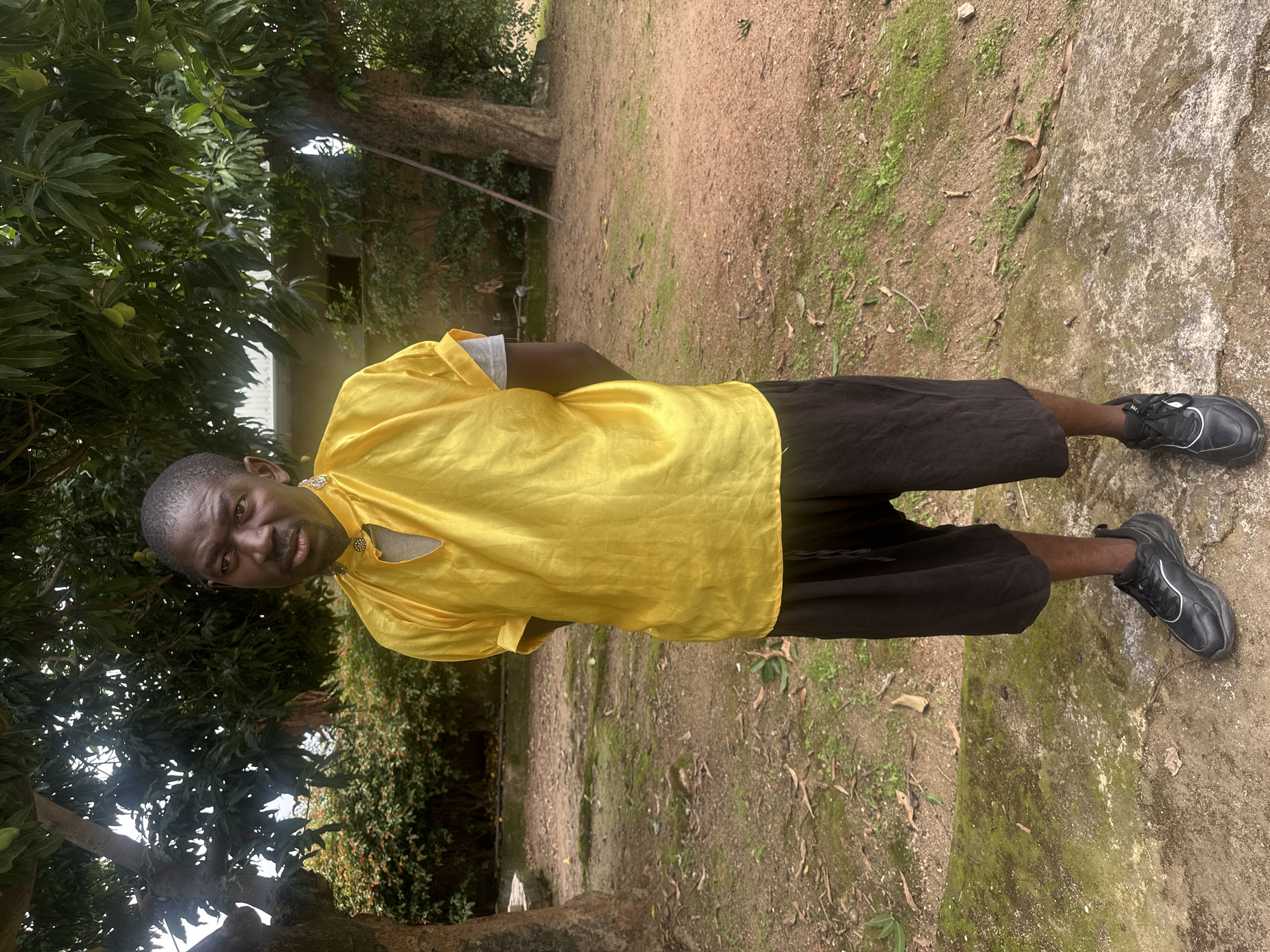 A Black man stands in a yard with scattered dirt and moss-covered ground. He wears a bright yellow tunic with a grey undershirt and black shorts, paired with black sneakers. The man is looking forward with his hands behind his back. Behind him, trees and bushes are visible, with a hint of a building in the background. The scene appears to be outdoors, possibly in a residential area of Jos, Nigeria, given the presence of mango trees and the typical architecture.