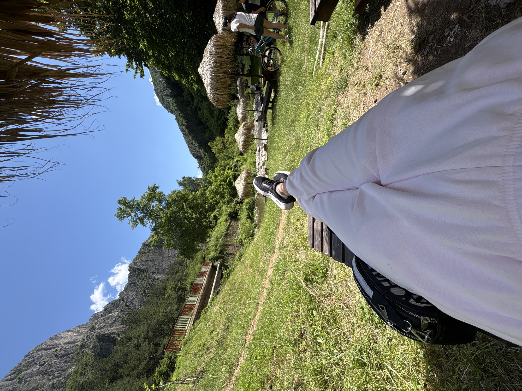 A first-person perspective shot captures a person relaxing on a wooden bench in a lush green park area. Their legs, clad in light pink pants, are extended forward, with their feet, wearing black and white sneakers, resting on the bench. A black bag with silver hardware is placed to the left of their legs.
The environment features a vibrant green lawn interspersed with patches of dry grass and scattered twigs. In the mid-ground, several thatched umbrella-like structures provide shade. A light blue bicycle stands near one of these umbrellas, with a person wearing a white t-shirt and denim shorts standing beside it. Another person, dressed in a dark shirt, is visible further back.
Beyond the park area, a shallow river flows, bordered by rocks and more greenery. A red and white railing runs along a raised path on a grassy slope to the left, which leads up to a sheer, rocky mountain face. The mountain is partly covered with dark green trees. The sky is a clear, bright blue with a few scattered white clouds. The scene is bathed in bright sunlight, suggesting it is a warm, sunny day. The location is identified as Zgorigrad, Bulgaria.