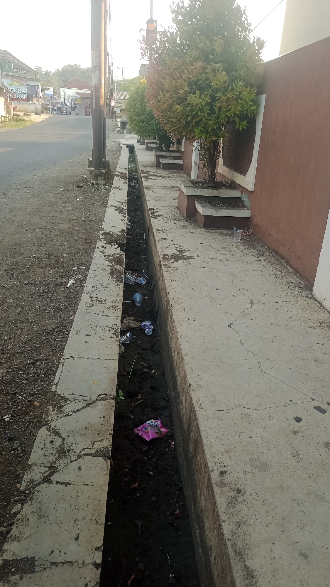 The image shows a street scene in Karyamandala, Indonesia, with a concrete drainage ditch running alongside the road. The ditch is filled with dark soil and debris, including several plastic bottles and a crumpled pink packet. On the sidewalk next to the ditch, there are potted plants with green and reddish foliage, as well as a brown-colored wall. The road appears to be asphalt, and there are some buildings and utility poles visible in the background, suggesting an urban or semi-urban environment. The sky is bright and appears to be early morning or late afternoon, given the soft lighting.