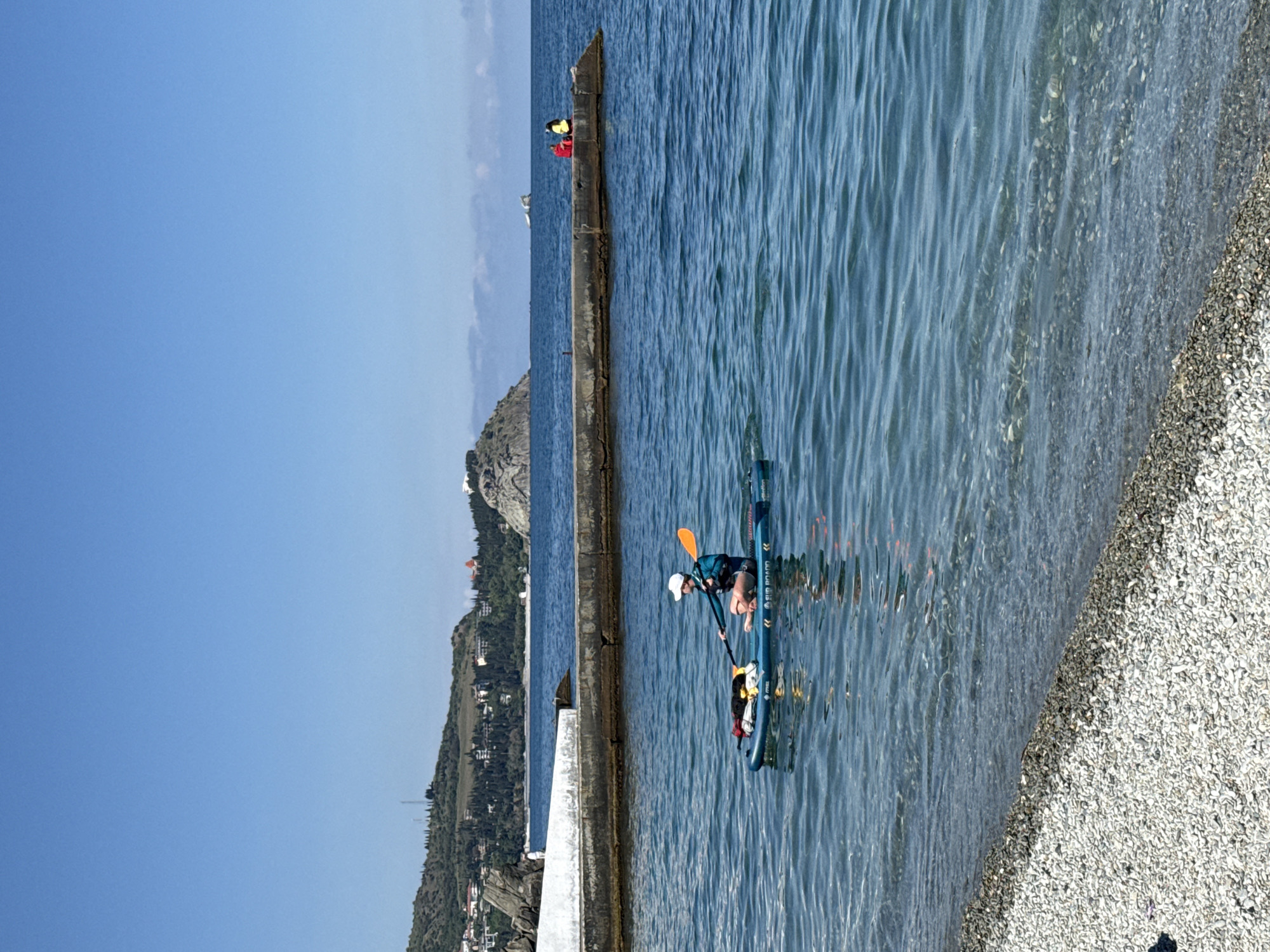 A sideways shot shows a person kneeling on a paddle board in the ocean in Unknown, Unknown. The person is wearing a hat and a blue and black top. There is a pile of bags on the front of the board. A paddle is lying down beside the person. The water is wavy, and there is a shoreline on the right. On the left, there is a concrete wall and a mountainous area in the background. There are a few people sitting on the concrete wall. The sky is clear and blue.