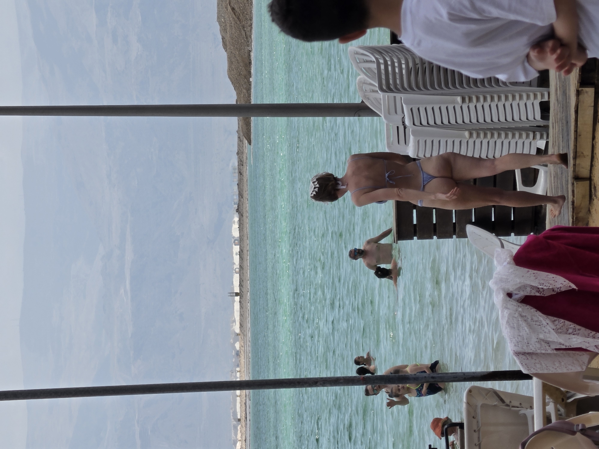 The image depicts a scene at the Dead Sea in Tamar Regional Council, Israel. The Dead Sea is in the background. In the foreground there is a metal support pole. The water has a distinct light-green-blue hue. Several people are in the water, some swimming and some wading. A woman is stepping onto a wooden deck from the water. A stack of white plastic chairs are stacked on the dock. A person is standing on the dock next to the chairs. A pink and white blanket is on the edge of the dock.