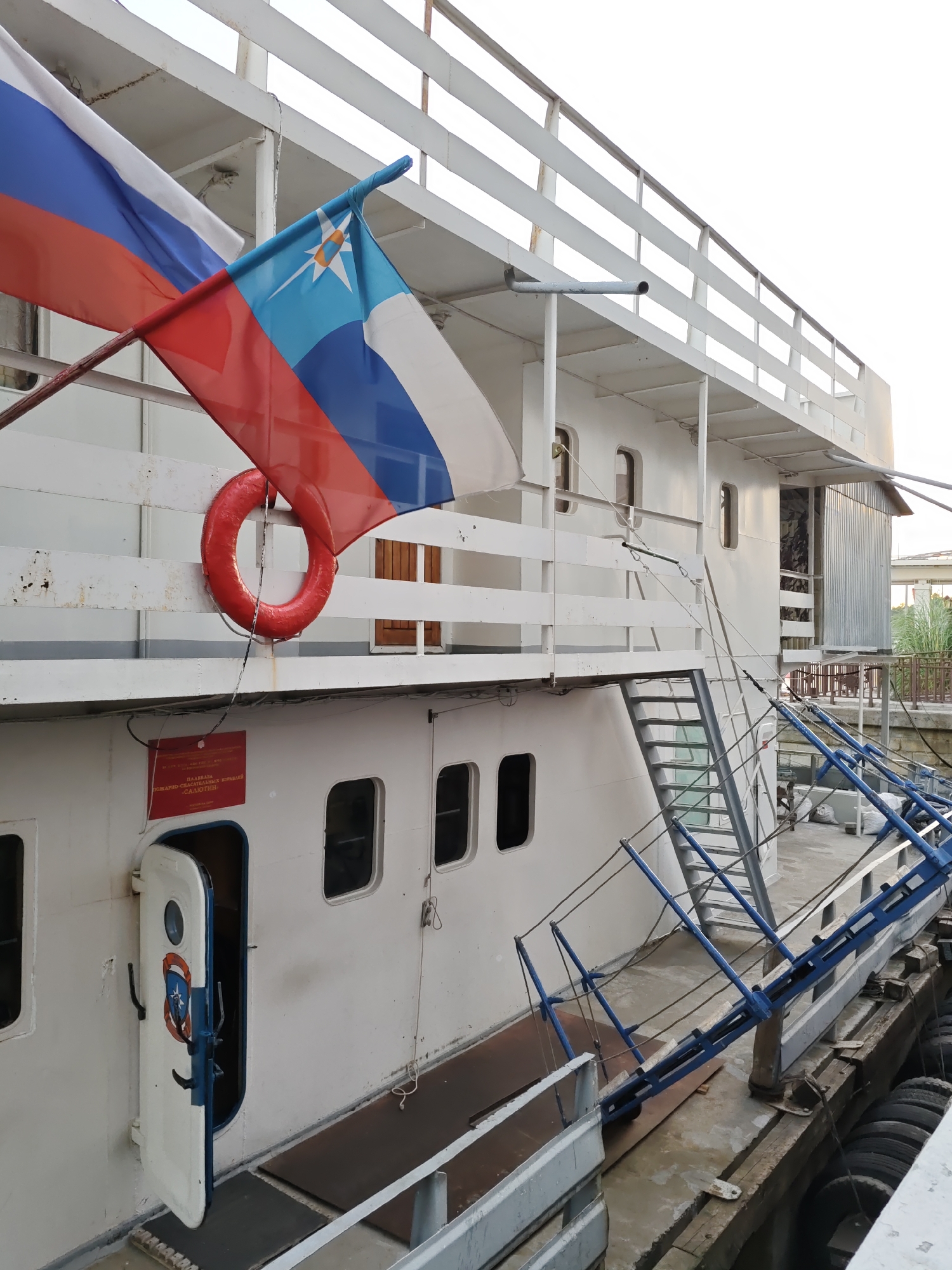 The image depicts a white ship docked on the embankment of the Don River in Rostov-on-Don, Russia. Two flags are prominently displayed on the ship's deck: the Russian flag and another flag with a blue background, a white star with an orange center, and white stripes. A red life preserver ring is attached to the railing. The ship has several windows and a white railing along its deck. A blue metal ramp extends from the ship to the dock. An open white door with a circular window and an emblem is visible on the side of the ship. A red sign with Russian text is also affixed to the ship's hull. The scene appears to be a still, outdoor environment with no people visible.
