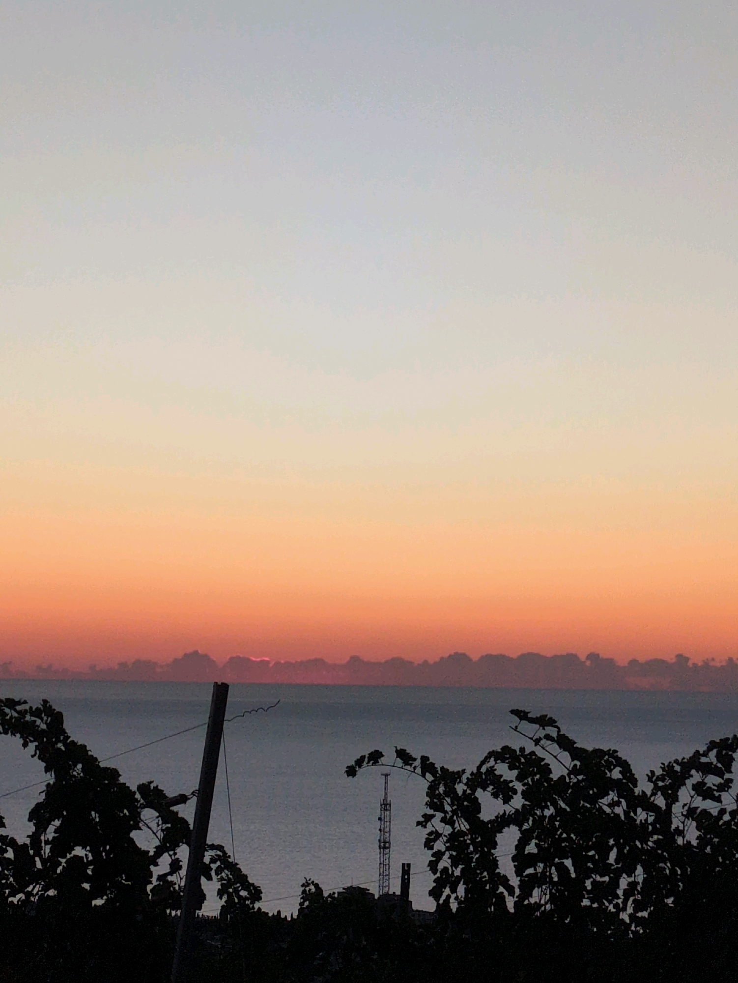 A silhouetted vineyard in the foreground overlooks a calm body of water. The sky transitions from a pale blue at the top to a warm orange, with a thin band of clouds just above the horizon. A sliver of the sun is visible peeking through the clouds. A wooden post with a wire is planted in the ground to the left of the frame, and a radio tower can be seen in the distance to the right. The overall impression is one of a serene sunrise or sunset.