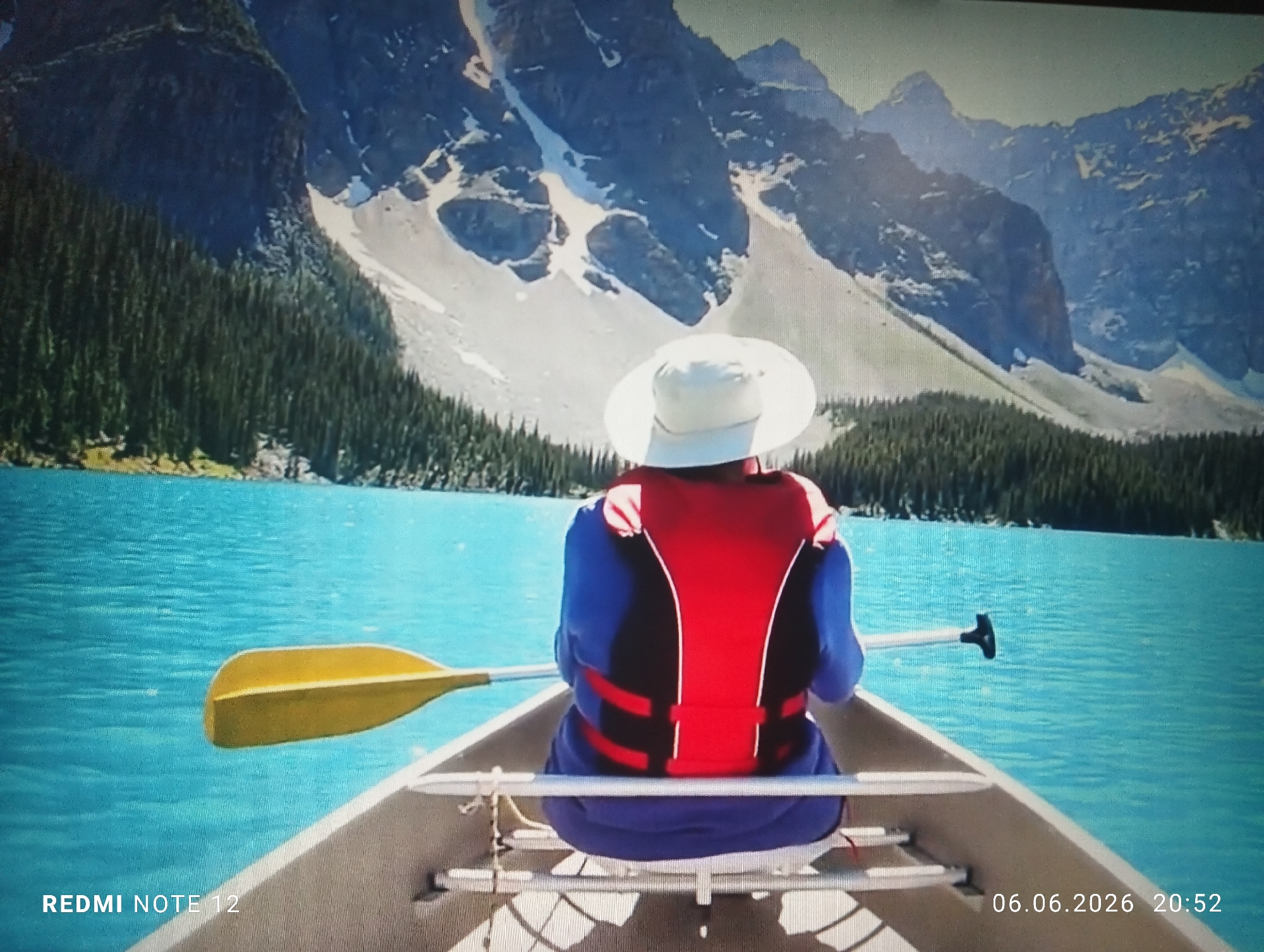 A single individual is observed from behind, seated in a light-colored canoe on a calm, turquoise-blue lake. The person wears a white wide-brimmed hat, a blue long-sleeved garment, and a red and black life vest. A yellow paddle is positioned horizontally across the canoe on the left, while the handle of another paddle is partially visible on the right.
The background features majestic, rugged mountains with prominent snow patches and dark rock faces. Dense coniferous forests line the foothills of the mountains and the shores of the lake. The sky above the mountains appears bright with a slight haze.
Text overlays are present: "REDMI NOTE 12" is visible in the bottom left corner, and "06.06.2026 20:52" is in the bottom right corner.
The scene depicts a high-altitude glacial lake surrounded by snow-capped mountains and dense coniferous forests, a landscape inconsistent with the city of Burgas, Bulgaria, which is a coastal city on the Black Sea. The geographic location strongly resembles regions within the Canadian Rockies.