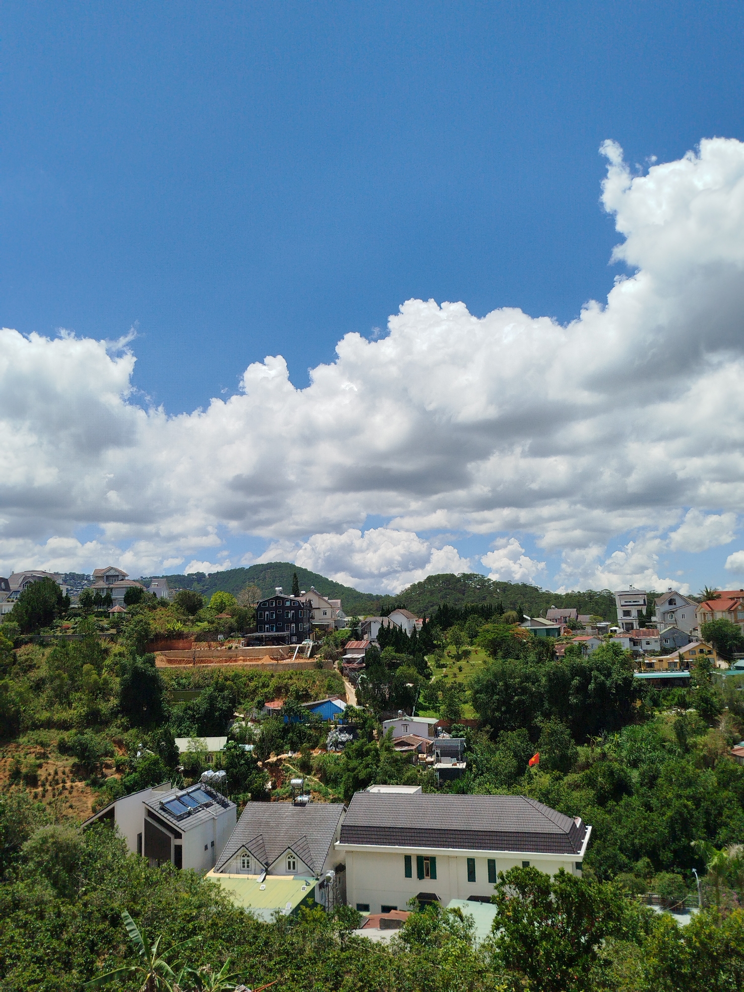 The image shows an elevated view in Da Lat, Vietnam, looking over a landscape of rolling hills covered in dense green vegetation and scattered residential buildings.
The sky is predominantly blue, partially obscured by cumulus clouds.
The buildings vary in style, some modern with solar panels on the roofs, and others with traditional designs, constructed with varying materials like tile roofs and concrete walls.
A construction site is visible on one of the hillsides, indicating ongoing development.
There are no people visible in the image. An orange Vietnamese flag is amidst the foliage.