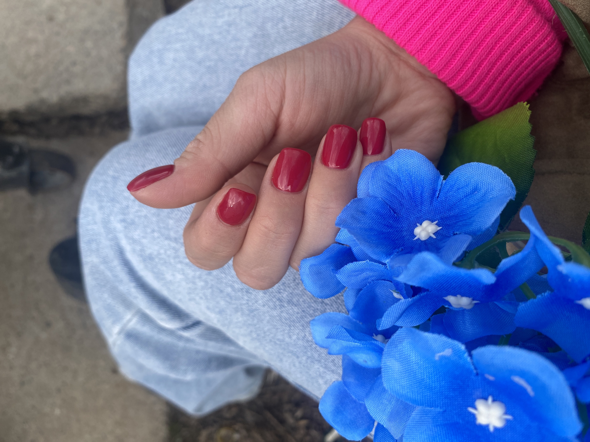 A hand with red nail polish is positioned next to a bouquet of artificial blue flowers with white centers. The hand rests on a pair of light blue denim pants. A bright pink sweater sleeve is partially visible near the wrist. A small portion of the ground and what appears to be a shoe can be seen in the background. The image was taken in the city of Chaikovsky, Russia.