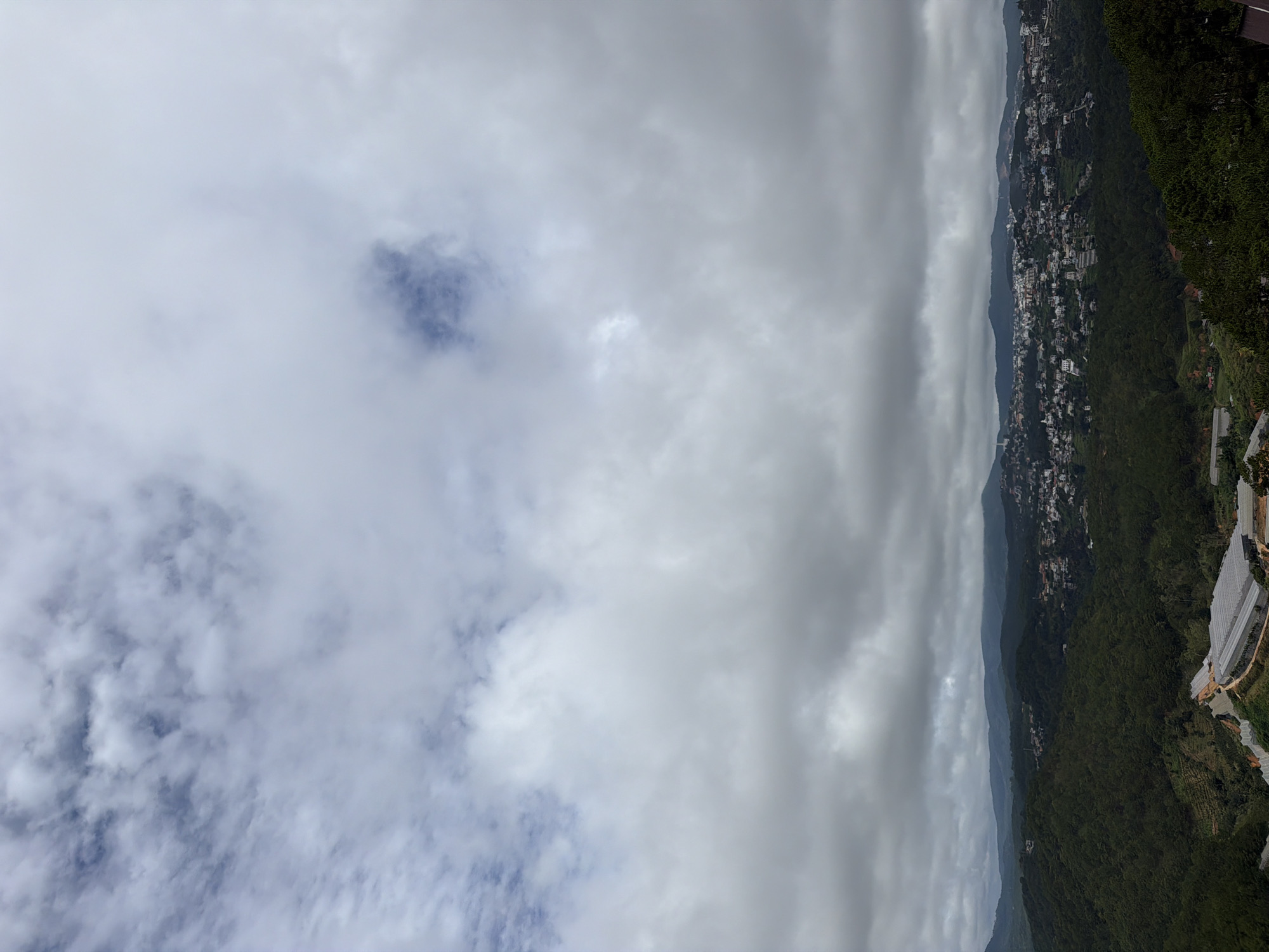 The image presents a panoramic view of a densely forested hillside with a town nestled in the valley. The sky is overcast with dramatic, layered clouds, ranging from light gray wisps to darker, more solid masses. The clouds dominate the upper portion of the frame, with hints of blue sky visible through thinner patches.
The mid-ground features a sprawling town with numerous buildings, predominantly white and red-roofed, dispersed across the undulating terrain. The town appears to be situated on a slope, with structures cascading down the hillside. Some roads are visible, winding through the urban landscape.
The foreground is characterized by lush, dark green vegetation, forming a thick forest. Terraced agricultural areas and greenhouses with translucent roofs are visible on the slopes, indicating human activity and cultivation. The landscape is mountainous and hilly, with the town and forests extending into the distance, fading into a hazy horizon. The overall impression is one of a rural or semi-urban setting in a mountainous region, under a cloudy sky. The location is identified as Xuân Thành, Vietnam.