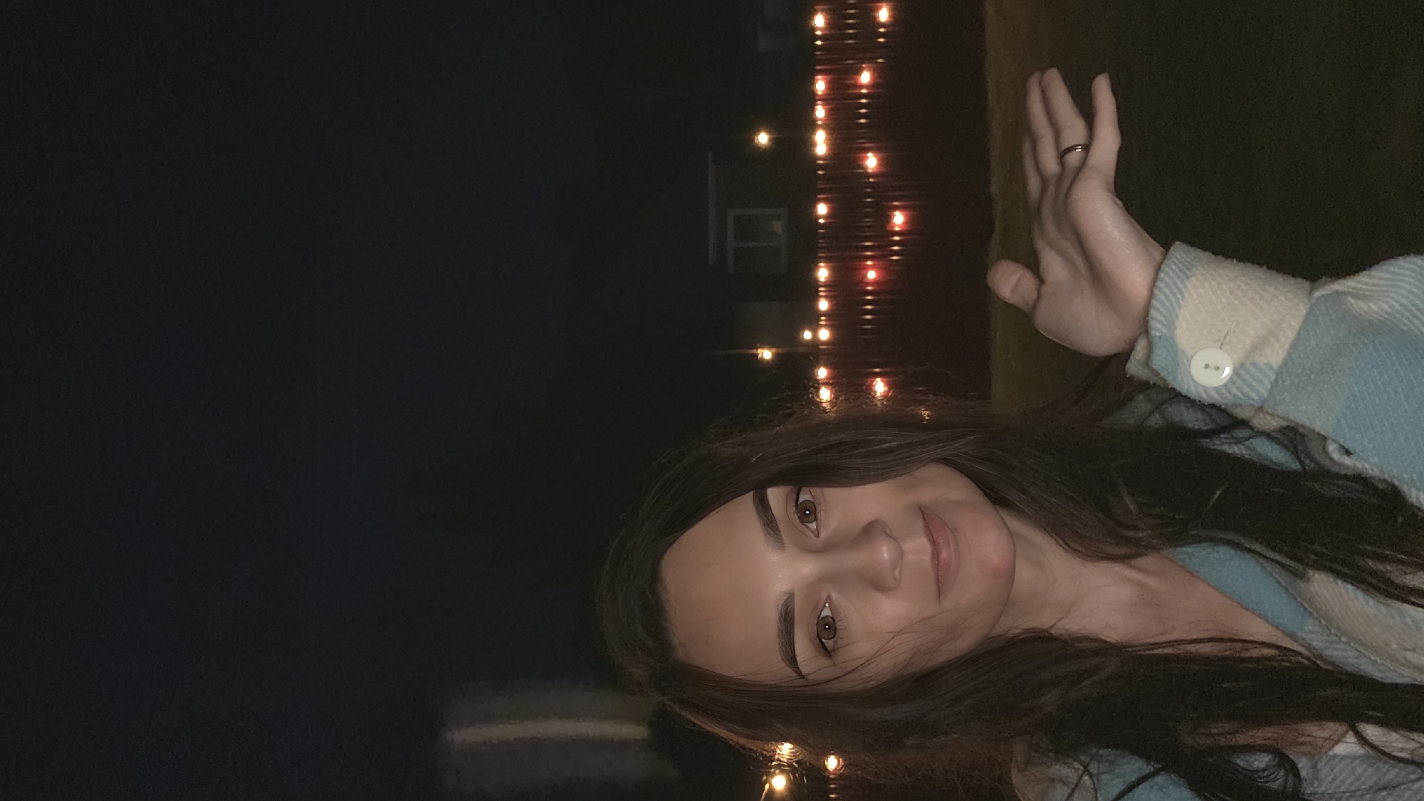 A woman with long, dark hair is pictured from the chest up, outdoors at night. She is looking directly at the camera with a slight smile. She is wearing a blue and white plaid shirt with a white button on the cuff. Her left hand is raised in front of her, palm up, with a ring on her ring finger. In the background, there is a fence with red and orange lights, and a dark, blurry building. The sky is dark. The location is Unknown, Unknown.