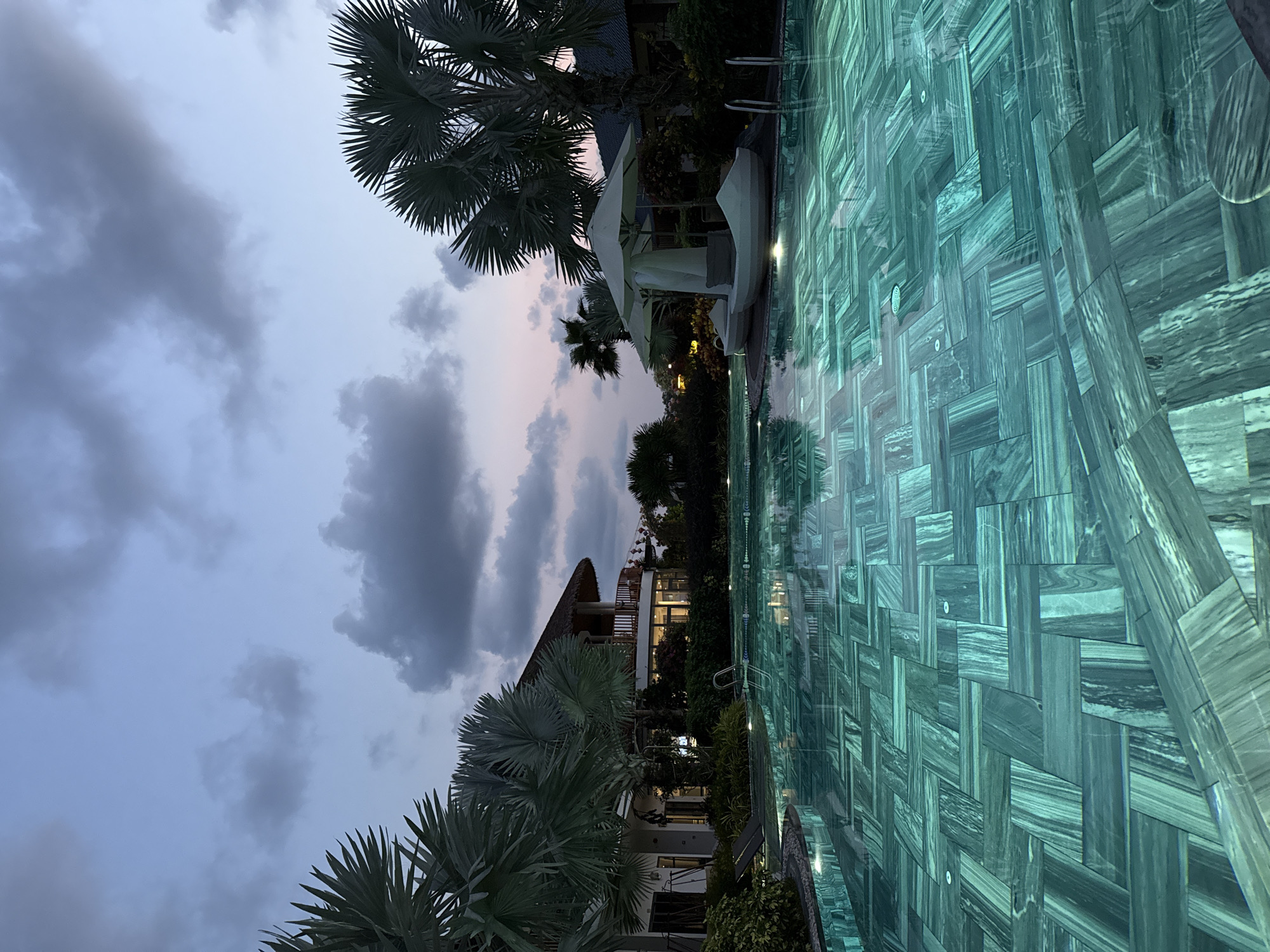 The media file captures a serene resort scene at dusk, likely in Phú Quốc, Vietnam. The primary subject is a large swimming pool that stretches across the right side of the image, illuminated by submerged lights. Its bottom is covered with distinctive green and blue marble-like tiles arranged in an intricate basketweave or herringbone pattern, giving the water a vibrant, reflective quality.
Along the pool's edge, several white, cushioned daybeds or loungers are visible, accompanied by large white parasols, indicating a luxurious and relaxing environment. Lush, fanned-leaf palm trees are scattered throughout the scene, framing the view and reinforcing the tropical setting.
In the background and mid-ground on the left, multi-story resort buildings are illuminated with warm interior lights. One building features a prominent thatched roof, a common architectural style in tropical regions. The sky, seen above the buildings and trees, is a soft twilight blend of light blue and grey, with scattered darker clouds. Subtle hints of pink and purple are visible near the horizon, confirming the time of day as dusk or just after sunset.
The scene conveys a peaceful and tranquil atmosphere, suggestive of a high-end vacation destination. There are no people visible, nor any discernable text. The unique pool tiling and the blend of natural and artificial light are notable details.