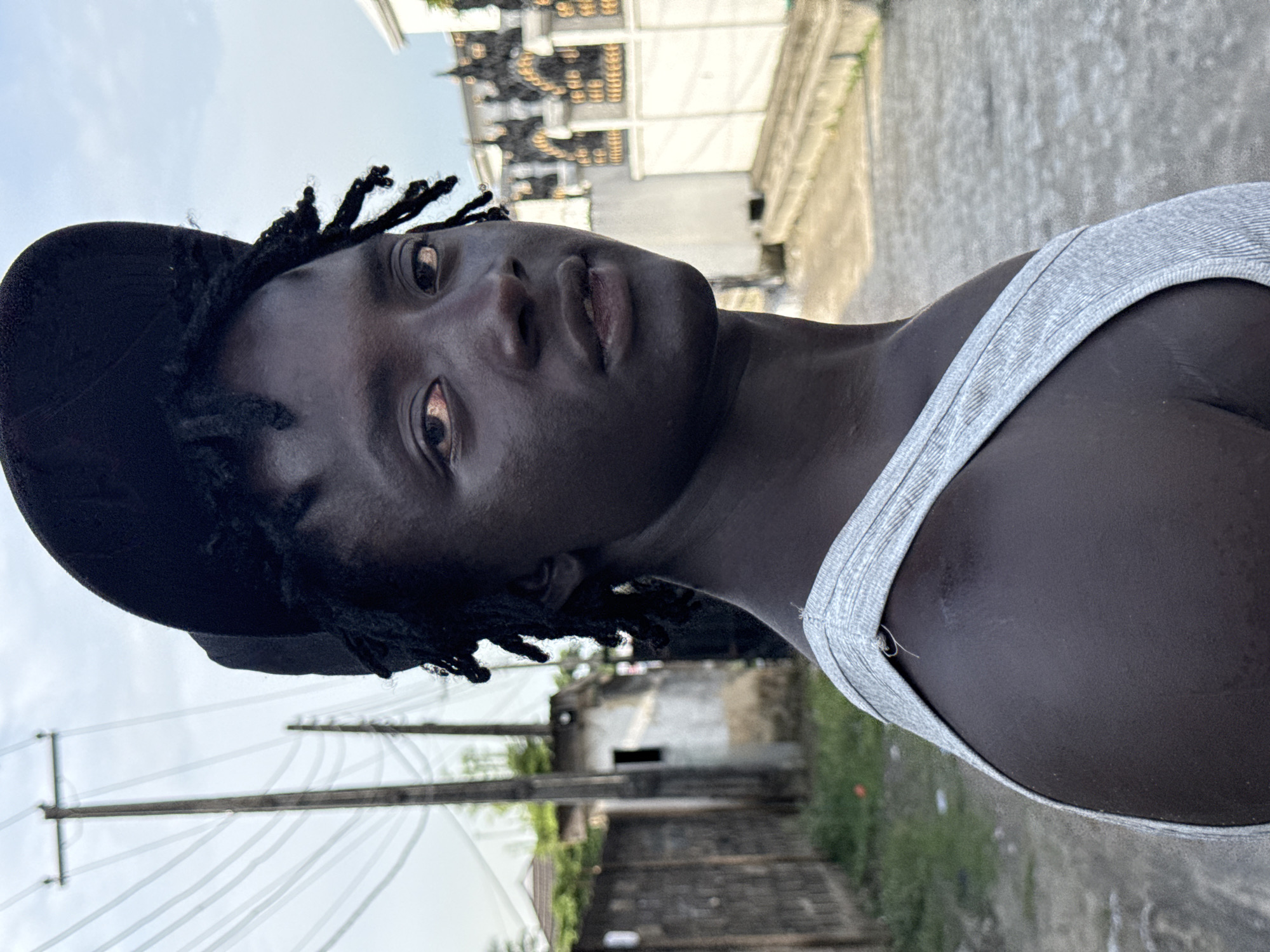 A low-angle, portrait-oriented photograph shows a Black person wearing a grey tank top and a black baseball cap, looking at the camera. Their hair is in dreadlocks. The background reveals a cityscape with visible power lines and aged buildings. The location is likely Ado, Nigeria, based on the prompt. The overall impression is casual and direct.