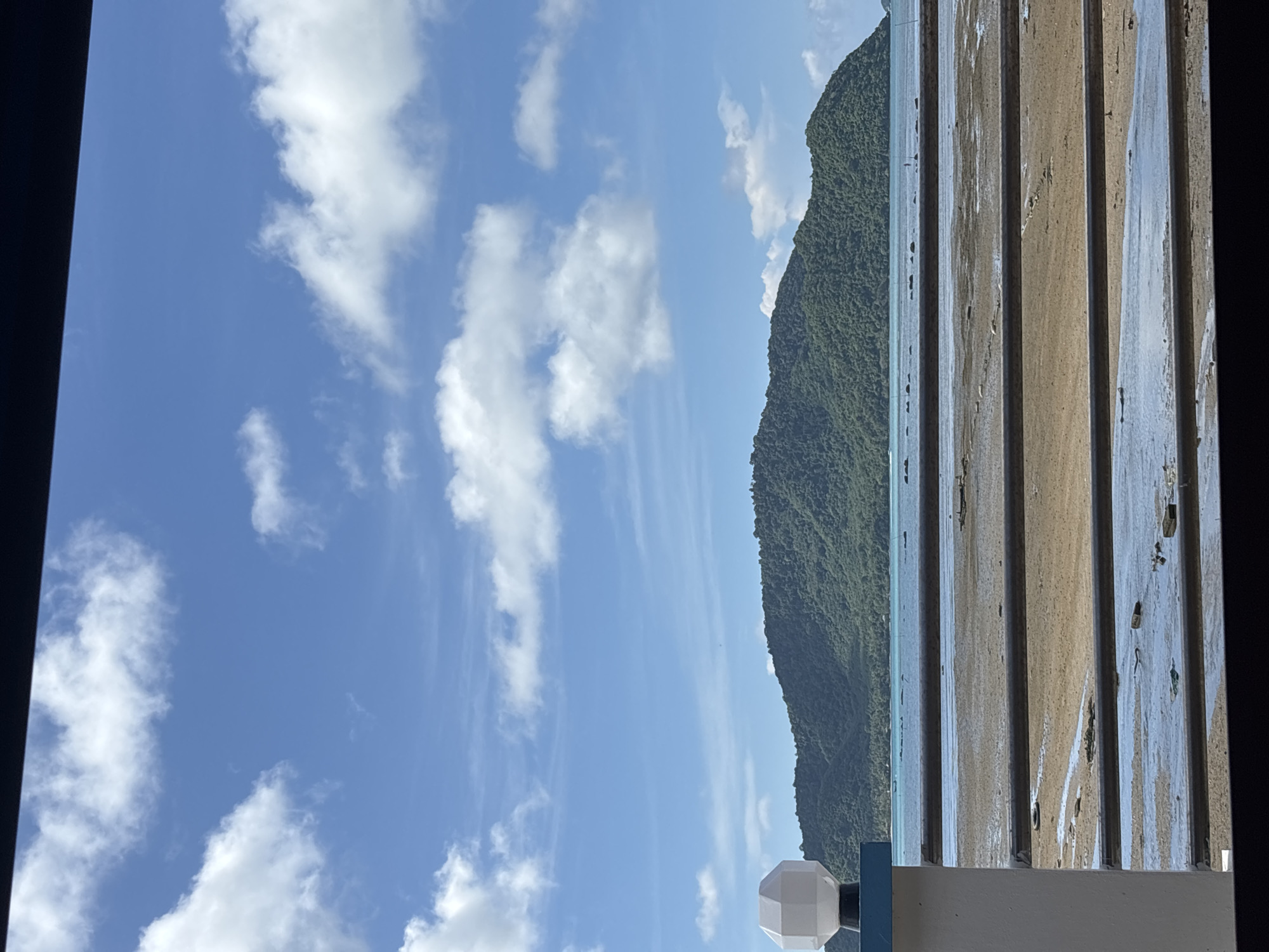 The image captures a scenic view of a tropical island with lush green vegetation, set against a bright blue sky dotted with fluffy white clouds. In the foreground, a white, geometric lamp fixture is mounted on a blue and white post, partially obscuring the view of a muddy beach with shallow water and scattered debris. Horizontal dark bars, likely part of a railing or fence, are visible across the lower half of the image, dividing the view of the beach and the distant island. The water appears calm, reflecting the sky, and a thin strip of sand is exposed. The overall impression is one of a peaceful coastal landscape in Rawai, Thailand, likely during daylight hours.