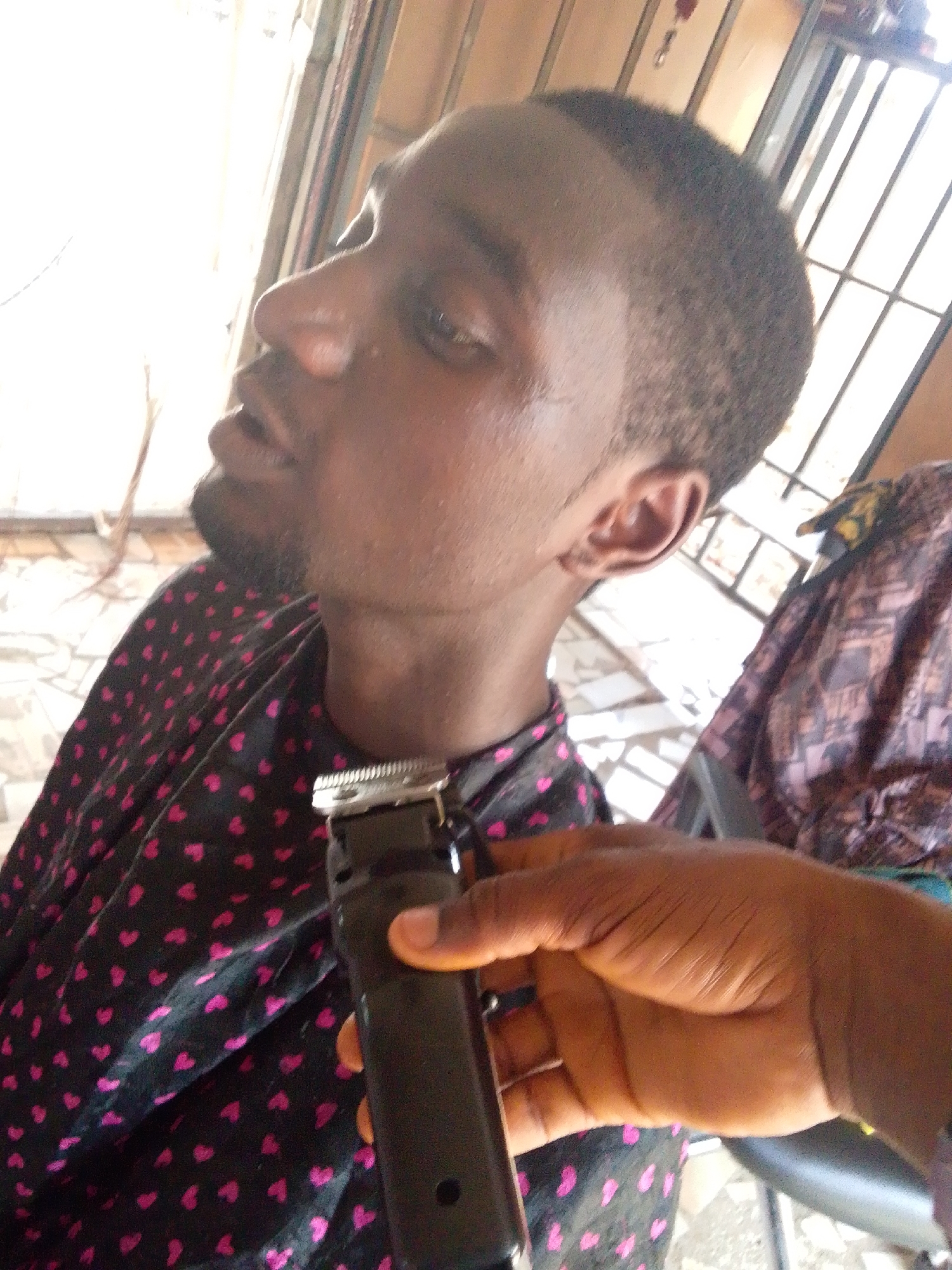 A dark-skinned man is seated and facing toward the left of the frame while receiving a haircut. The man has a short haircut with a clean line along the hairline. A small beard is also present. The individual is wearing a black shirt patterned with small pink hearts. A hand, holding black hair clippers, is in front of the man's neck. The setting appears to be indoors, possibly a home, with a window visible in the background. The location is in the city of Unknown, Unknown.