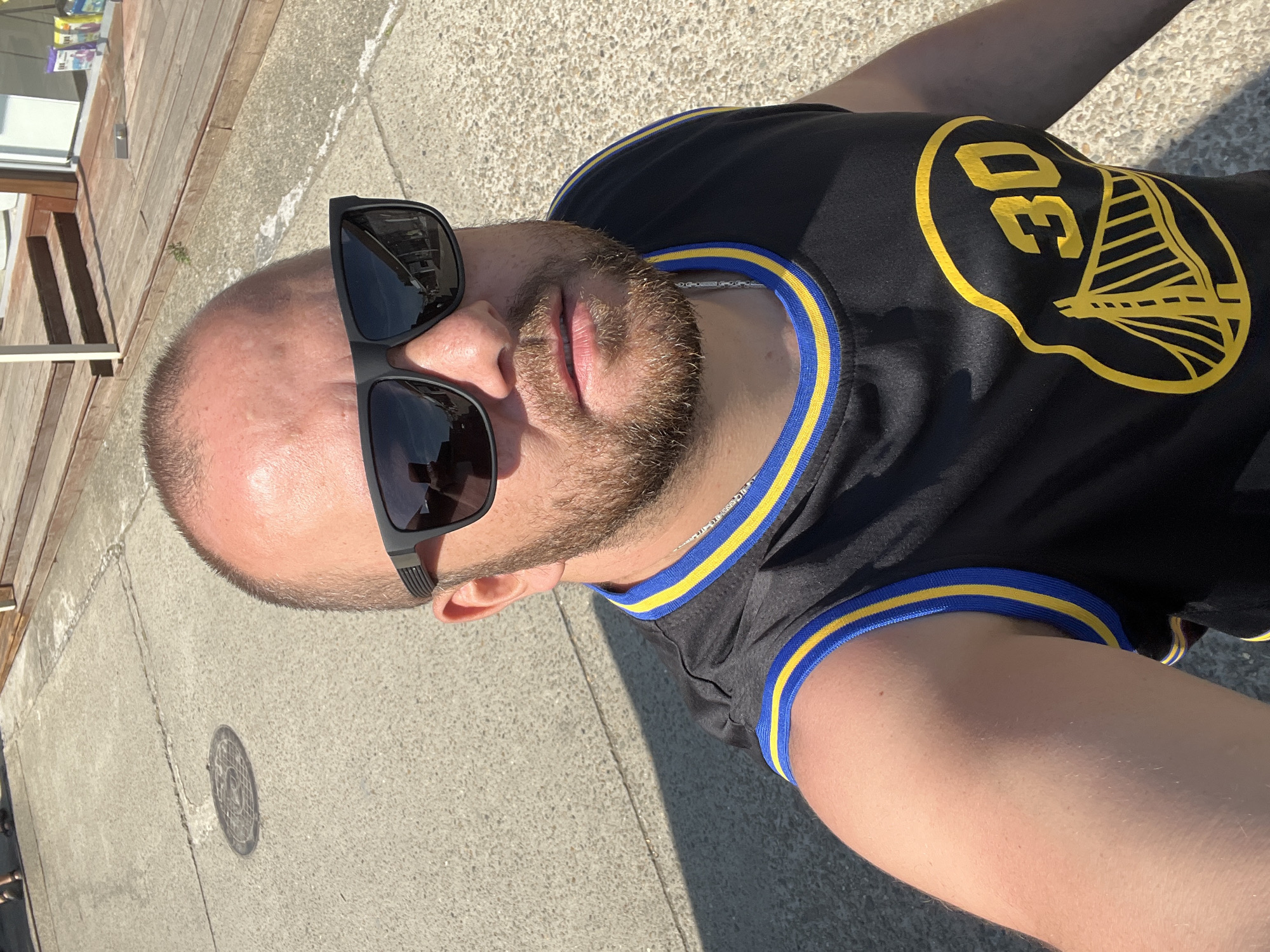 A male individual, appearing to be in his 30s to 40s, is depicted in a high-angle selfie taken outdoors in the city of Sochi, Russia. He has a fair complexion, a receding hairline with short dark hair, and a neatly trimmed brown beard. He is wearing dark, rectangular sunglasses that reflect a distant street or interior, and a black sleeveless athletic jersey with yellow and blue striped trim around the neck and armholes. The jersey's front features a prominent yellow graphic comprising the number "30" and a stylized depiction of the Golden Gate Bridge, consistent with the Golden State Warriors NBA team logo. A silver-toned chain necklace is visible below the jersey's neckline. The background consists of a light grey concrete ground surface, which exhibits cracks and a circular metal manhole cover to the upper left. To the upper right, part of a building is visible, featuring a light-colored facade, a wooden deck or steps, and a storefront displaying various packaged goods behind glass. The scene is brightly illuminated by direct sunlight, casting distinct shadows on the ground from the individual's head and shoulders.