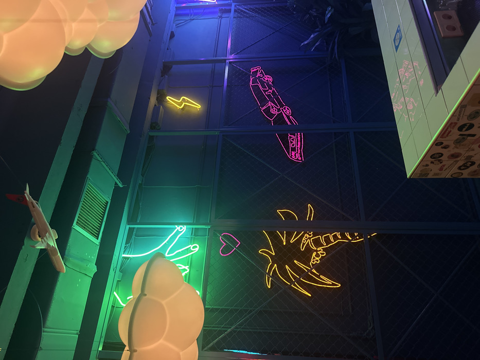The image displays the interior of Freedome Retro Future Bar, located at ul. "Bratya Miladinovi" 26, 1000 Sofia Center, Sofia, Bulgaria. The environment is dark with a prominent display of various neon light installations against dark metal grid partitions.
From the dark ceiling, a white model airplane with a red tail is suspended. Below it, an exposed air duct glows green. Multiple large, white, multi-lobed cloud-shaped light fixtures are visible, with one prominent in the upper left. The wall partitions feature distinct neon art: a green outline of a hand making a peace sign, a pink outline of a heart, a yellow outline of a palm tree, a yellow lightning bolt, and a pink outline of a car resembling a DeLorean, with the word "Freedome" illuminated in pink neon beneath it. In the lower right foreground, a section of a bar counter is visible, featuring a light green tiled top and a side adorned with a collage of small labels or stickers. Dark foliage is present behind the bar counter. No individuals or specific actions are depicted within the scene.