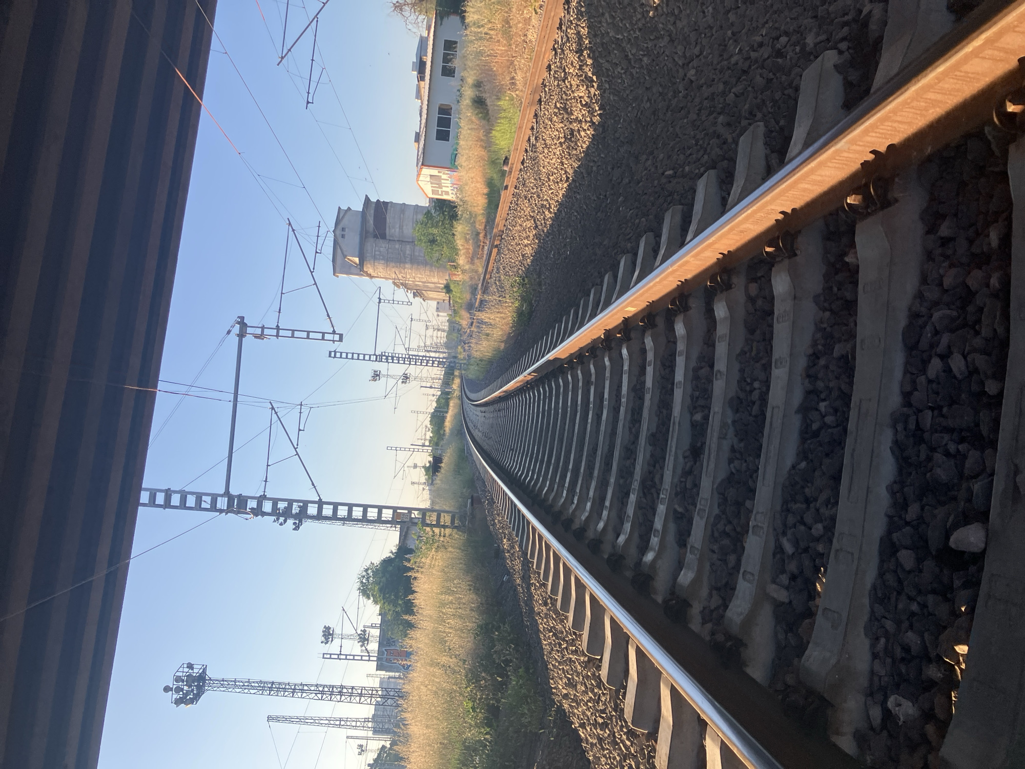 The image depicts a railway track in Plovdiv, Bulgaria, curving into the distance. The tracks are flanked by gravel and grassy vegetation. Electrical poles and overhead wires run parallel to the tracks. In the background, a building complex is visible. To the left, a structure is seen, its scale unclear. The sky is blue. There is no visible human activity.