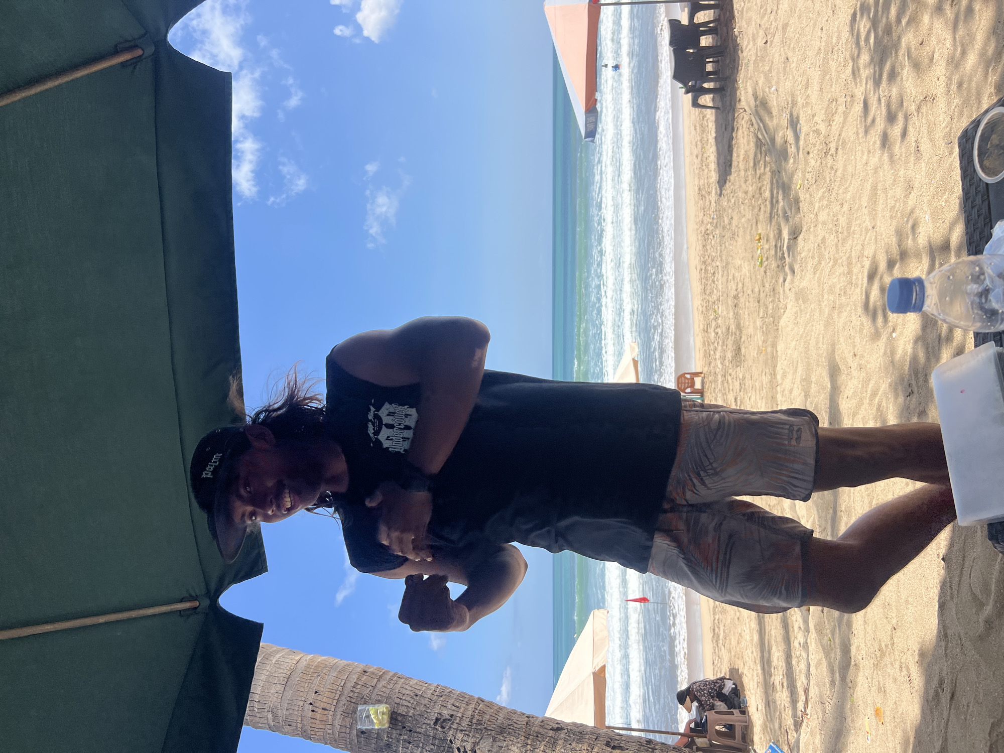 A man stands on a beach in Kuta, Indonesia, under a large green umbrella. He wears a black sleeveless t-shirt with a graphic print and patterned shorts. He is looking to his left and has his arms crossed over his chest. In the background, the ocean with waves is visible, along with a few beach umbrellas and chairs. The sand is light brown, and a palm tree trunk is on the left side of the image. A clear plastic water bottle with a blue cap is in the foreground on a table. The sky is blue with scattered white clouds.