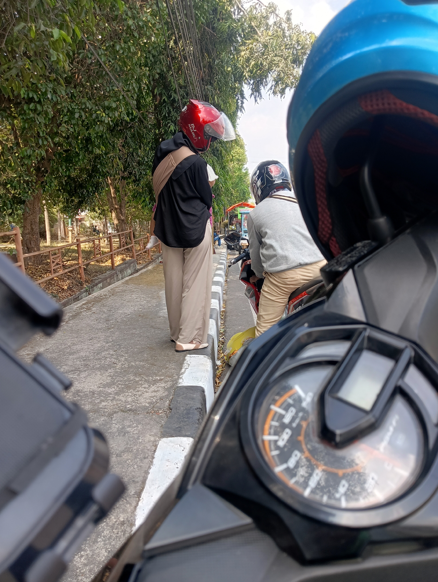 Two individuals on motorcycles are positioned on a paved road adjacent to a sidewalk with a concrete curb. The individual in the foreground, on the right, is wearing a blue helmet and is facing away from the camera, with their body partially visible. The motorcycle in the foreground is in focus and shows a black instrument panel with a circular speedometer. The second individual, further down the road and on the left, is wearing a red helmet and a black jacket over beige pants. They are standing next to their motorcycle and appear to be interacting with someone or something out of frame. The environment is characterized by trees lining the road, a wooden fence, and a cloudy sky. The scene is captured in Cijoho, Indonesia.