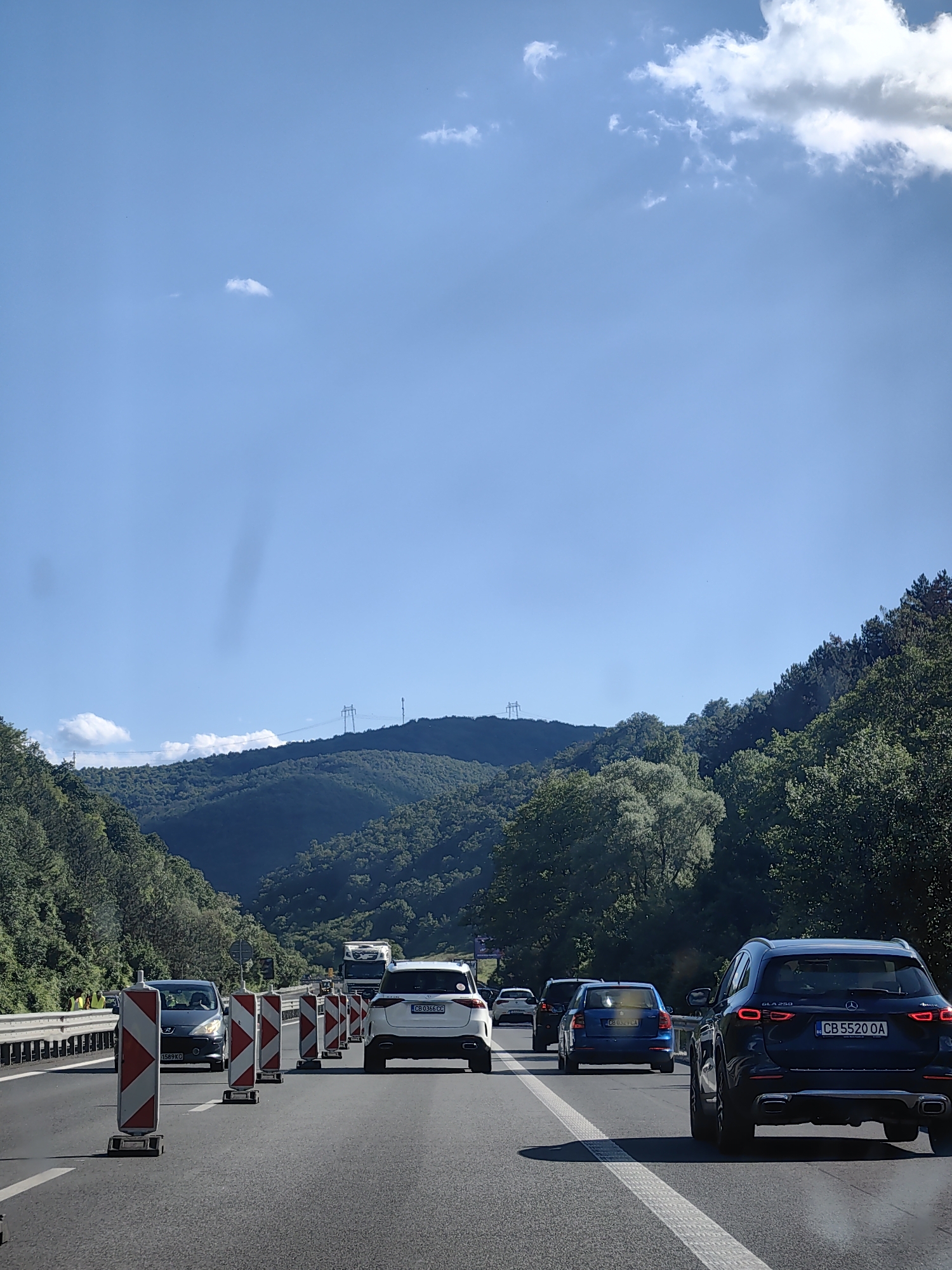 The image depicts a highway scene in Churek, Bulgaria, on a sunny day with a clear blue sky and scattered clouds. The highway is lined with trees on both sides, with rolling green hills in the background. Several cars are visible on the road, including a dark blue Mercedes-Benz GLA 250, a white SUV, a blue hatchback, and a dark-colored sedan. Traffic cones and barriers are set up on the right side of the road, indicating construction or maintenance work. Two individuals wearing yellow vests are standing near the barriers on the left side of the road, likely workers. Power lines and transmission towers are visible on the distant hills. The overall atmosphere suggests a typical day on a Bulgarian highway.