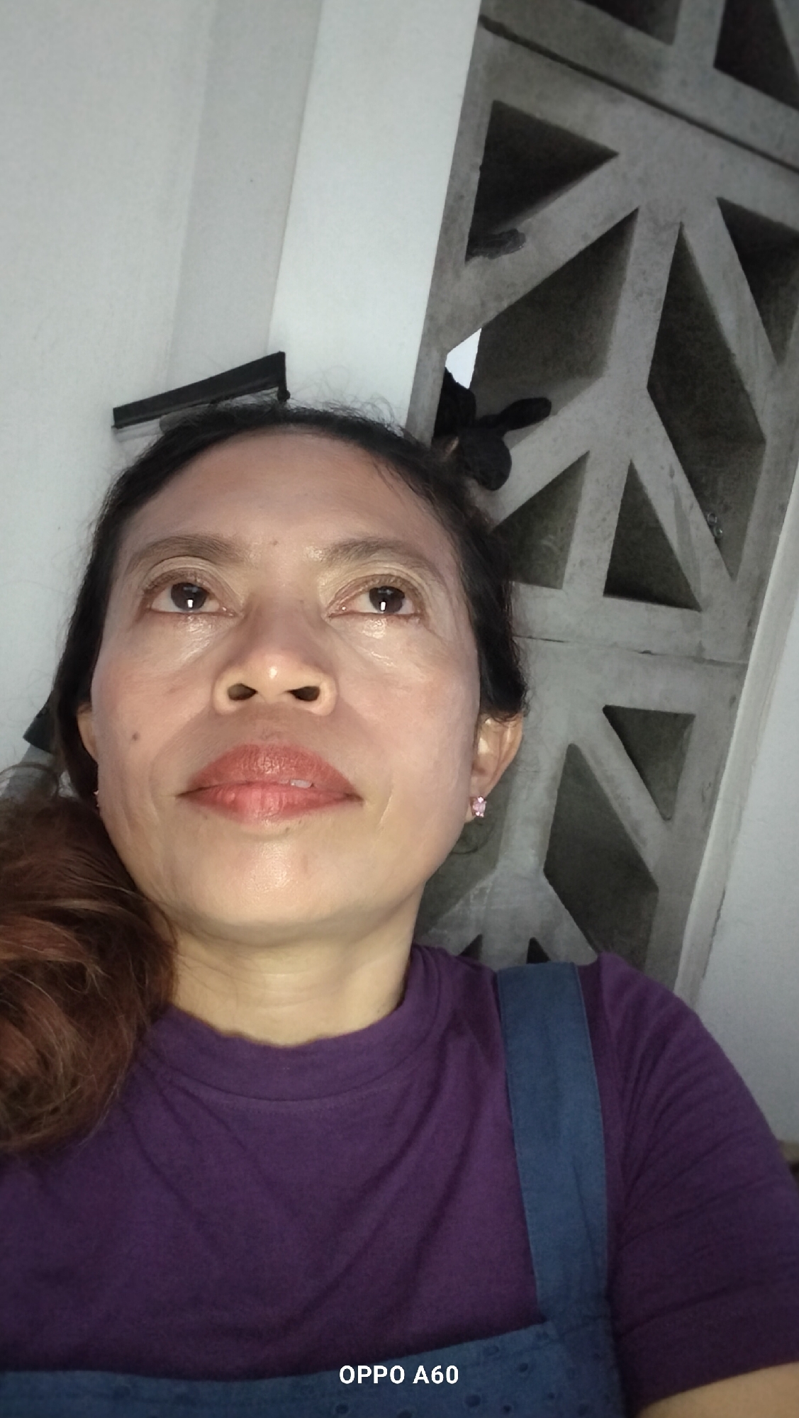 A middle-aged Indonesian woman is pictured in a close-up, head-and-shoulders shot, looking upwards and to her left. She has medium-toned skin, dark hair pulled back, and is wearing a purple t-shirt and a blue denim pinafore. A small pink earring is visible in her left ear. The background consists of a white wall and a gray, geometric concrete screen. The image appears to have been taken indoors, possibly in a residential setting in North Kuta, Indonesia.