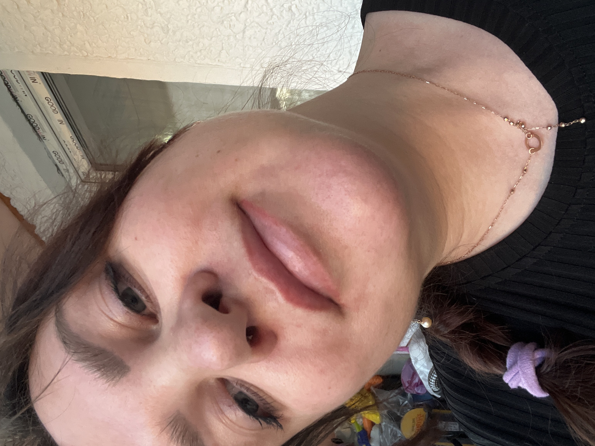 A close-up, slightly angled, indoor selfie shows a person with light skin, dark hair, and dark eyes. They are wearing a black top and a delicate gold necklace. Their hair is pulled back into pigtails with a purple scrunchie and a decorative hair pin. In the background, a window with white frames and lettering that says "GOOD IN" is visible, along with assorted items including wrappers and packaging materials. The location within the city of Noviy, Russia, cannot be determined from the image.