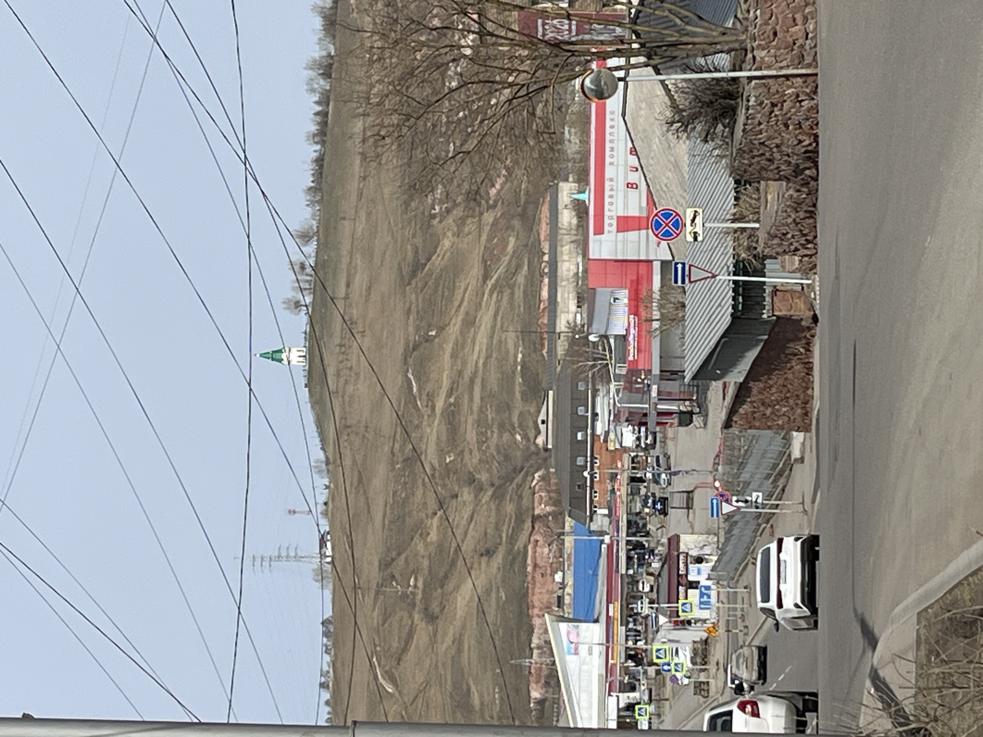 The image shows a street scene in an unknown city, possibly in Russia, based on the signage. The road is bordered by a sidewalk and various commercial buildings, including a large red and white building with the words "ТОРГОВЫЙ КОМПЛЕКС" on the facade. A large, barren hill dominates the left side of the frame, with a church tower visible at its crest. Several power lines intersect the view. Vehicles, including white cars, are parked or driving along the road. Street signs and other urban infrastructure are also present. The sky is overcast, and the overall atmosphere is somewhat bleak.