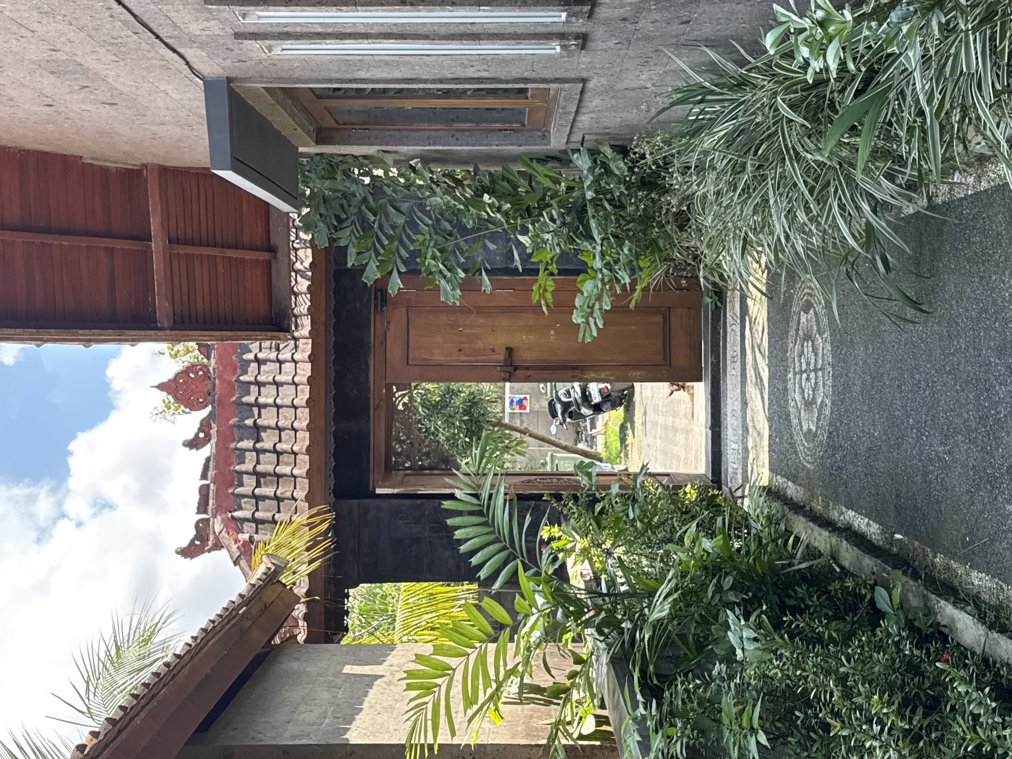 A perspective view shows an open, weathered wooden door leading from a courtyard-style structure out to a street in Kuta Selatan, Indonesia. The architecture of the building is a blend of traditional and modern elements with a tiled roof featuring decorative carvings. Lush greenery frames the door, creating a contrast between nature and built environment. Beyond the doorway, a street scene is visible with a motorbike and other vehicles suggesting urban activity. The courtyard features a patterned stone path, adding to the aesthetic appeal of the space. The scene gives an impression of a transition between a private, enclosed space and the public, bustling environment of Kuta Selatan.