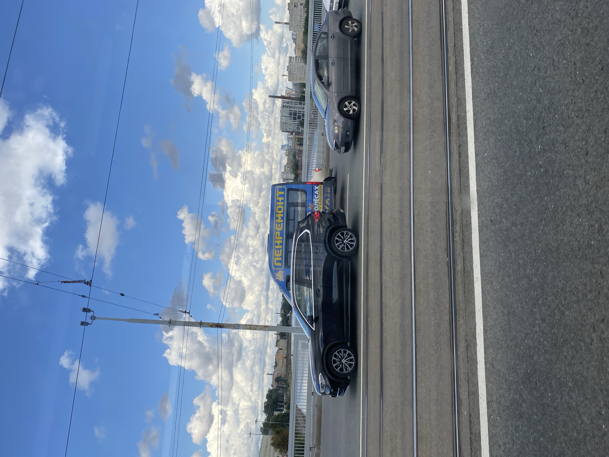 This is a medium shot, taken from a moving vehicle, looking out at a bridge over a body of water in Saint Petersburg, Russia. The sky is blue with scattered white clouds. A streetcar track runs across the bridge, and two cars are driving on the road. A blue van with Russian text on the side, reading "РЕМОНТ" (REMONTS), is parked on the side of the road. The van has images of cartoon characters on it. In the background, there are buildings and industrial structures. The bridge appears to be an urban bridge with a railing.