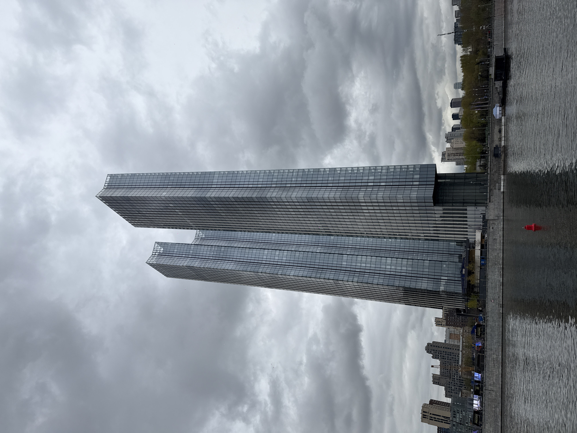 The image shows two modern, rectangular skyscrapers in Moscow, Russia, set against a cloudy sky. The buildings feature glass facades with horizontal lines. A body of water is visible on the right side of the frame, with a few smaller buildings lining the waterfront. The buildings are likely part of the Moscow International Business Center (Moscow City). The weather is overcast.