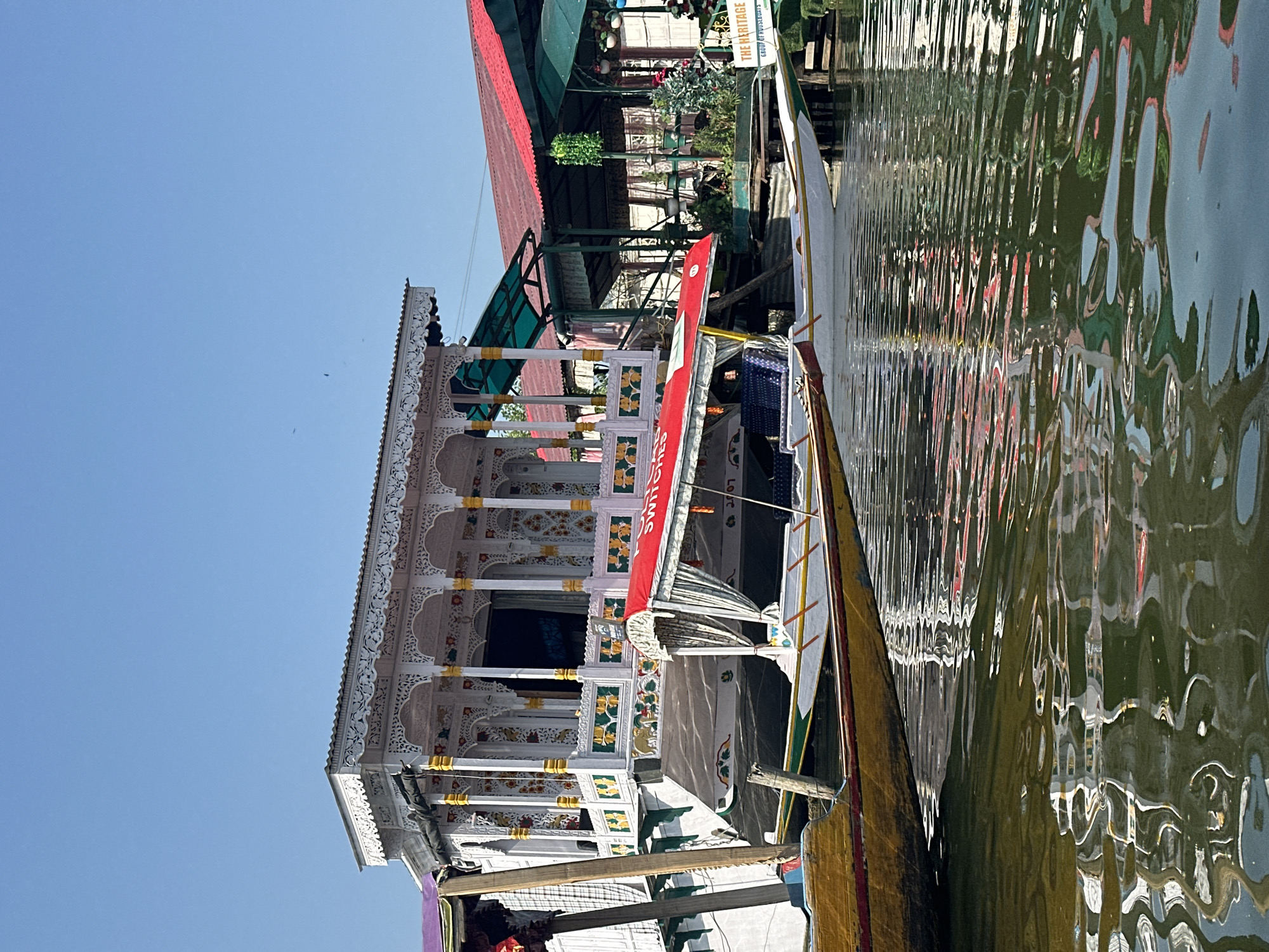 In Srinagar, India, on a clear, sunny day, a traditional Kashmiri houseboat is moored on a body of water, likely Dal Lake. The houseboat is elaborately decorated with intricate white carvings, gold accents, and colorful floral motifs. A smaller, vibrant red-covered boat is docked alongside it, with its curtains drawn. Other boats are visible in the background, along with what appears to be a covered seating area adorned with plants and hanging decorations, suggesting a tourist or hospitality setting. The water reflects the colorful structures, creating a shimmering, impressionistic effect. The scene is tranquil, depicting a typical day in the picturesque tourist landscape of Srinagar.