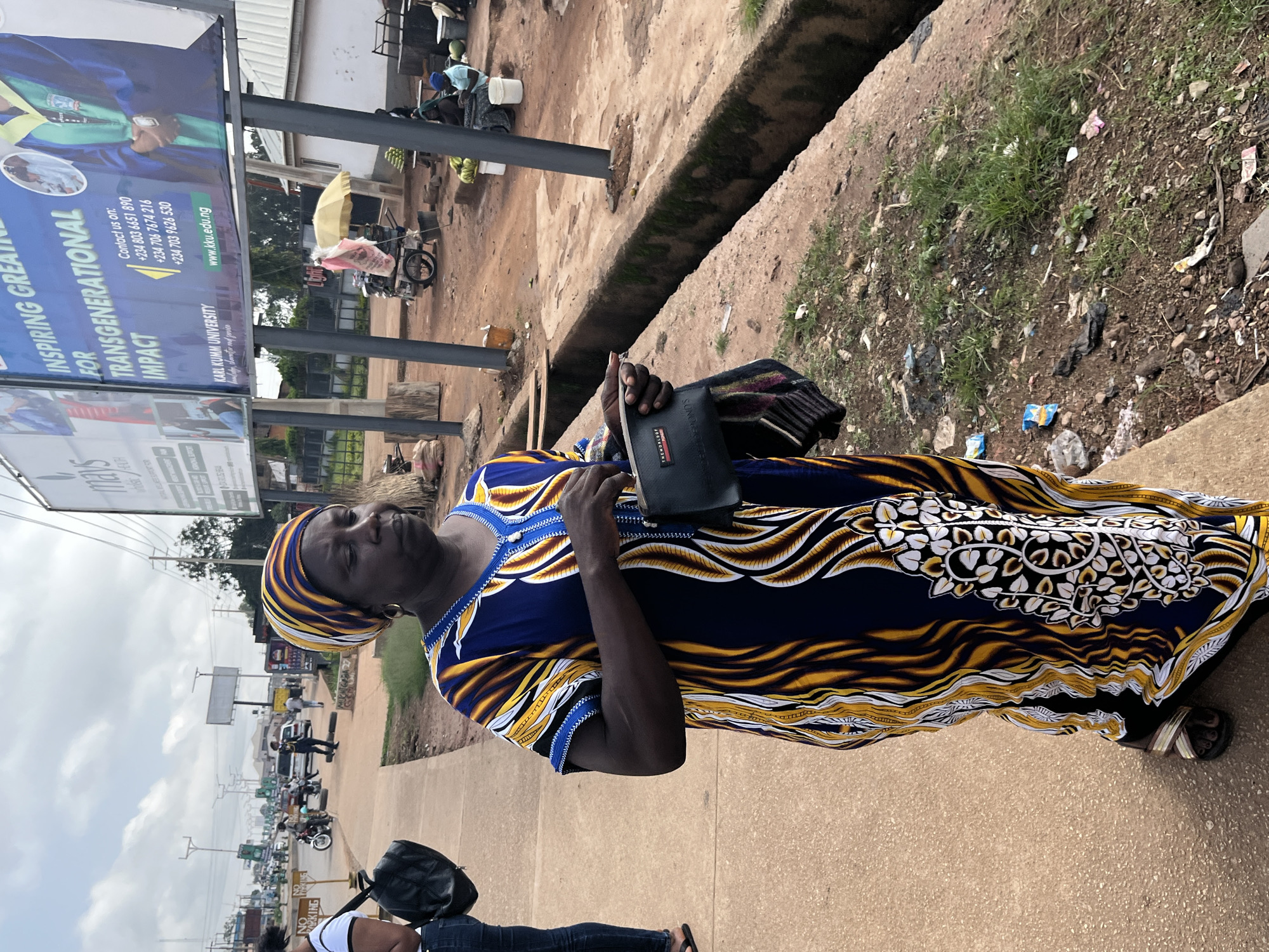 A medium shot captures a woman standing on a sidewalk next to a street in Gura Top, Nigeria. The woman, wearing a colorful blue and yellow patterned dress and a matching headwrap, is holding a black clutch purse. Her face is turned to the right, and she appears to be looking down. In the background, there's a busy street with cars and motorcycles, and a large billboard advertising Karl Kumm University. The sidewalk is concrete, and the ground next to it is dirt and grass, with some litter visible. The sky is overcast.