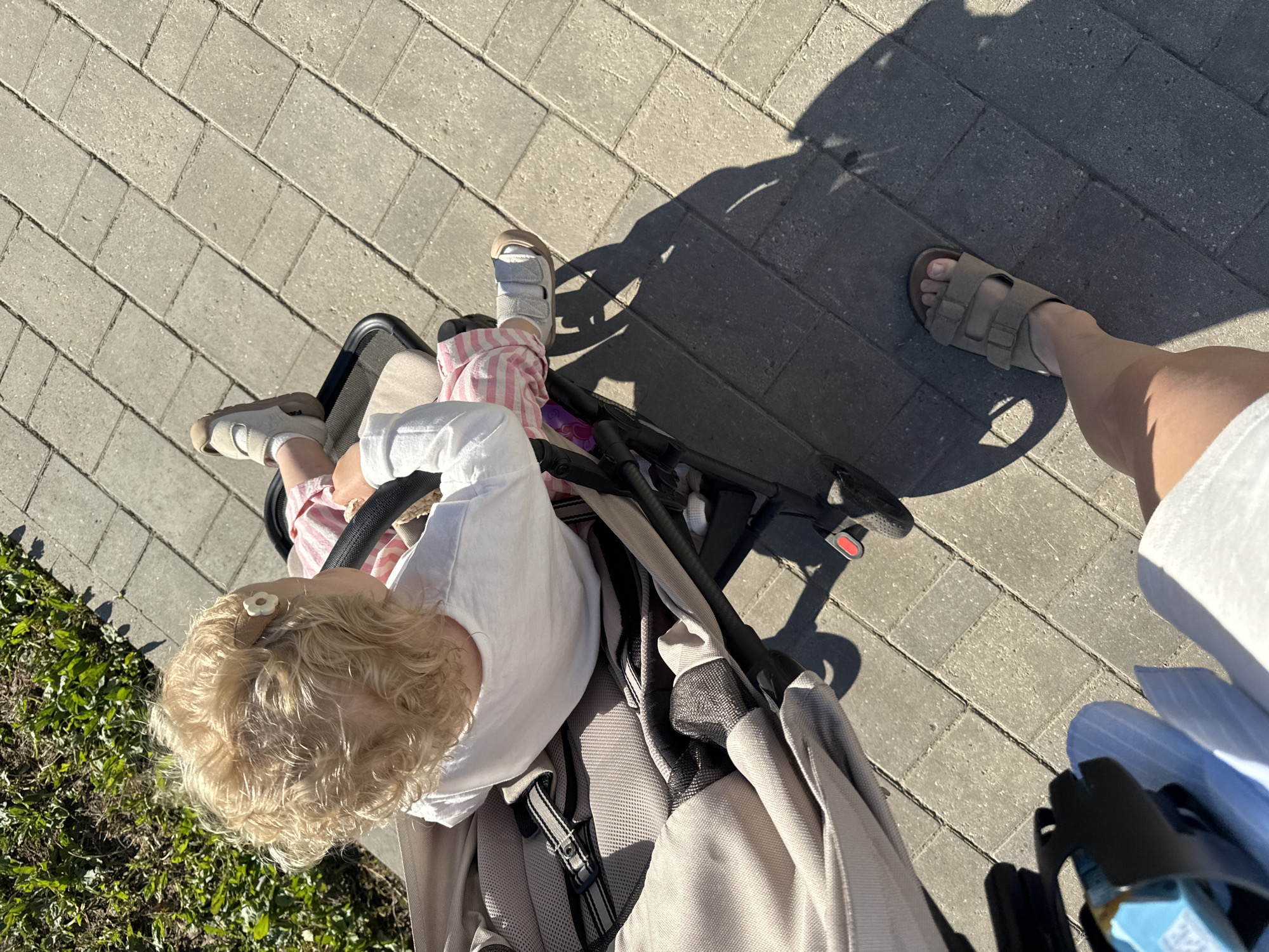 In a view from above, a toddler with blonde curly hair sits in a light-colored stroller on a sunlit paved pathway. The toddler wears a white long-sleeved shirt and pink and white striped shorts, with light-colored sandals on their feet. The stroller has a beige fabric seat and a black frame. To the right of the stroller, a person's sand-colored sandals and lower leg are visible, casting a shadow on the pavement. Green grass and vegetation are present to the upper left of the image. This scene is likely outdoors in a park or on a sidewalk in the city of Vladimir, Russia, indicated by the presence of outdoor paving stones and greenery typical of urban public spaces.