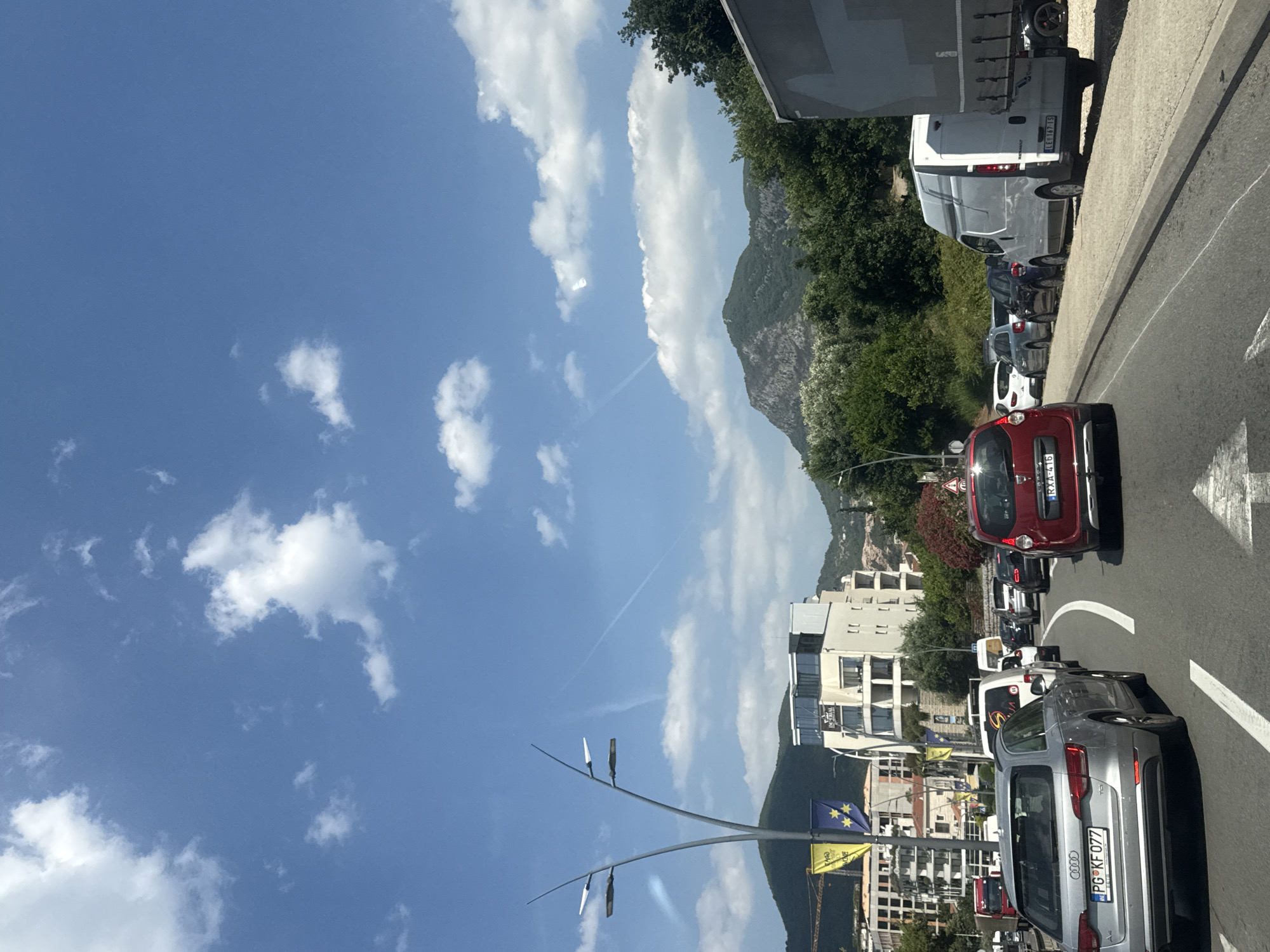 The image depicts a traffic jam on a sunny day in Budva, Montenegro. The foreground shows several cars stopped on a road, including a silver Audi A4 Avant with license plate PG KF 077. A red Dacia Sandero is in the lane to the right. Further ahead, a white van and a large truck are visible. In the background, there are buildings, lush green trees, and mountains under a blue sky with scattered white clouds. The presence of numerous cars indicates a busy urban environment.