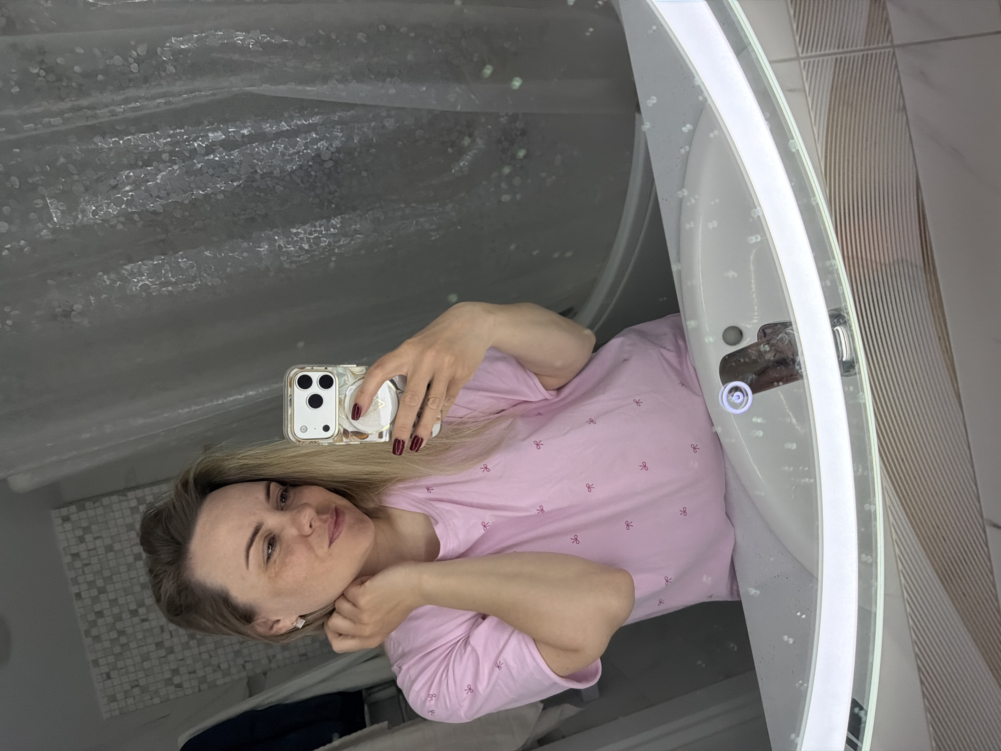 An adult female is depicted in a mirror selfie, captured within a bathroom located in Votkinsk, Russia. The subject, a fair-skinned woman with light blonde hair, displays light freckling on her cheeks and wears diamond-shaped earrings. She is dressed in a light pink short-sleeved t-shirt adorned with small, darker pink bow tie patterns. Her visible right hand, with dark red manicured nails, holds a light-colored smartphone encased in a patterned cover, which is positioned for the selfie. Her visible left hand is raised, with fingers resting near her jawline and hair. She gazes slightly to her left with a subtle smile. The bathroom environment includes a circular mirror featuring an illuminated LED border, positioned above a white sink equipped with a chrome single-lever faucet. Water droplets are evident on the mirror's surface, particularly in the lower portion near the sink. A wall section with small, light-colored mosaic tiles is visible behind the woman's reflected left side, and a textured, translucent shower curtain is visible on the reflected right side. Below the sink, a section of light-colored, horizontally ridged wall tiles is visible.
