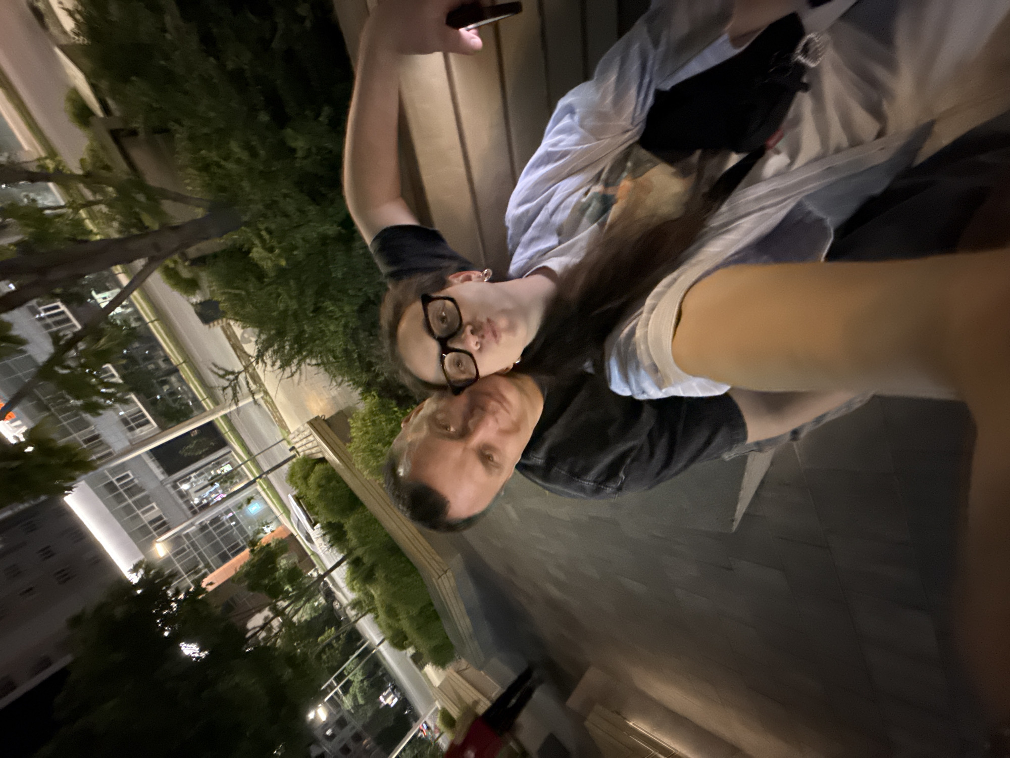 A nighttime selfie photograph shows two people, a man and a woman, seated outside in what appears to be an urban environment. The woman, wearing glasses and a light-colored top with a dark layer underneath, is holding the phone to take the picture. A man with short hair is next to her. Buildings are visible in the background, illuminated by street lights. The surroundings suggest a city environment with trees and street lamps along a sidewalk. The photograph was taken outdoors in Kazan, Russia, based on the street signs and architectural style that can be seen in the background.