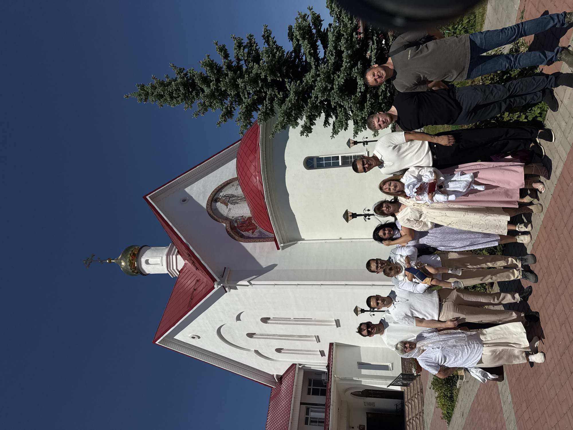A group of approximately 15 people stand in front of a white building with a red roof and a gold-domed tower. The building appears to be a church. The people are a mix of men, women, and children. Some of the women are wearing traditional dresses. The men are wearing casual clothing. The group is positioned on a brick sidewalk. A green tree is behind the building. The sky is blue. The image appears to be taken in the city of Unknown, Unknown.