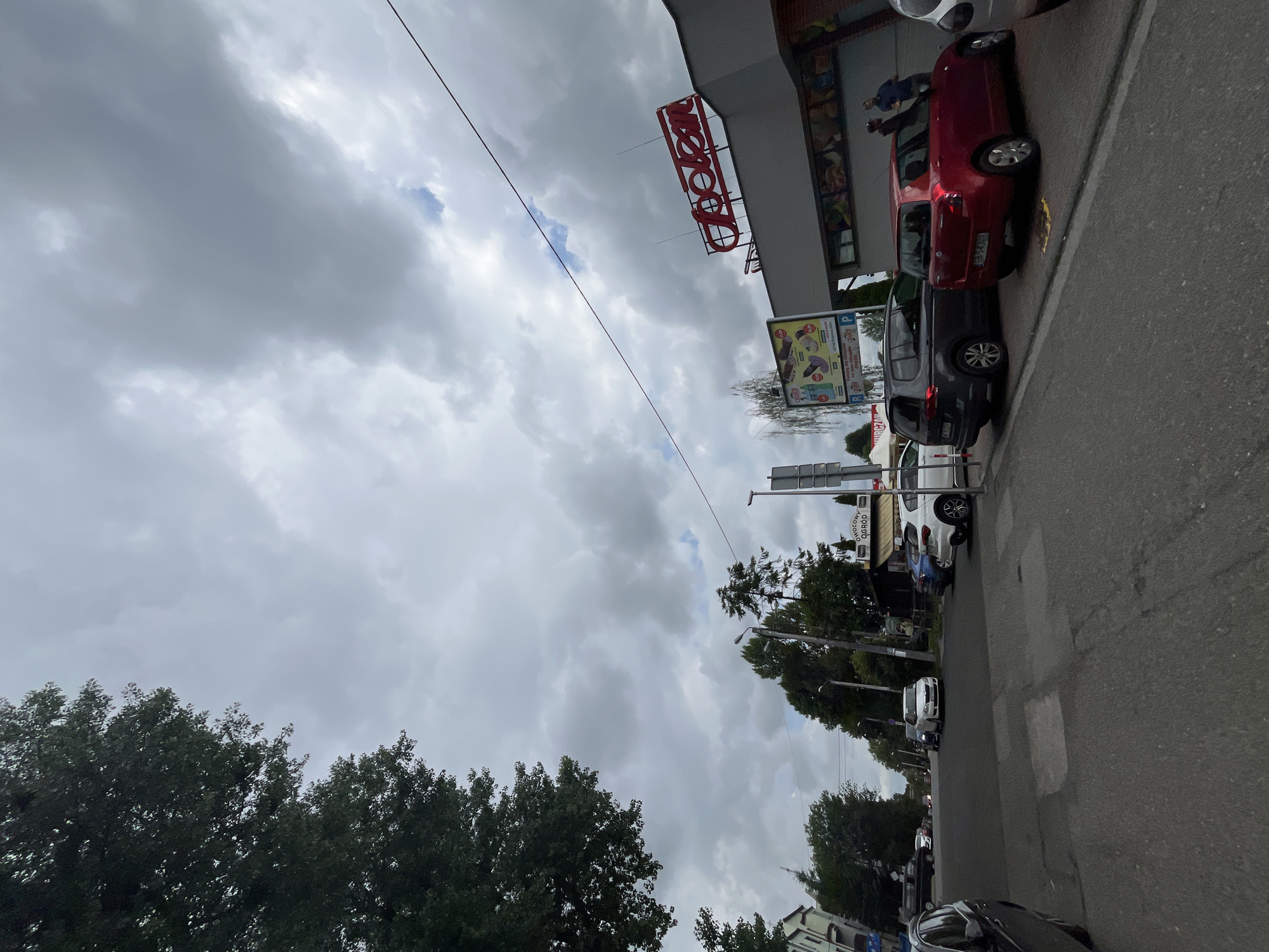 The image shows a street scene in an urban environment under a cloudy sky. On the right side of the street, a building with a red neon sign that reads "Biedronka" is visible. A red hatchback car is parked in front of the building, with two men standing near it. Several other cars are parked along the street, including a dark gray SUV and a white hatchback. Trees line the left side of the street, with their branches extending towards the sky. The road is paved with asphalt, and there are some painted markings on it. A billboard advertising various products is mounted on the side of the building. The overall atmosphere is overcast and somewhat subdued.