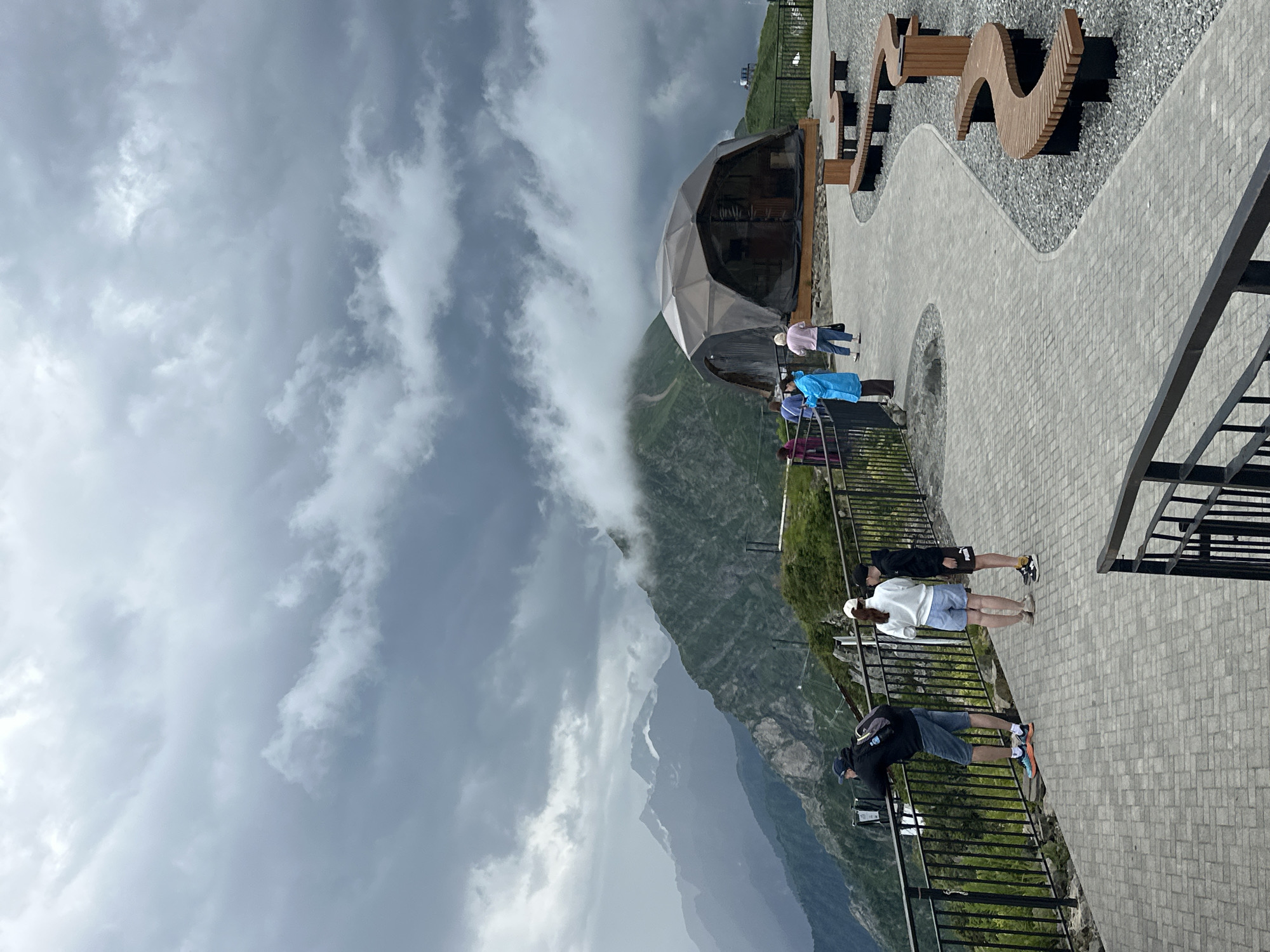 This is a photograph taken from a scenic overlook in Estosadok, Russia, with a dramatic, overcast sky and mountain landscape. In the foreground, a paved walkway curves to the right, bordered by a dark metal railing. Several people are visible on the walkway. One man in a black hoodie and blue shorts leans on the railing, looking out at the view. To his right, a woman in a white top and blue shorts and a person in a black hoodie and shorts stand together. Further along the path, a group of people, including one wearing a bright blue raincoat, are walking towards a large, geodesic dome structure. This dome, with a dark, translucent material covering its frame, is situated on a grassy knoll. Behind the dome, the rugged, green slopes of mountains rise, partially obscured by thick, grey clouds that also dominate the sky. The mountains in the distance are sharp and jagged, with some appearing snow-capped. Unique, S-shaped wooden benches are placed on the gravel to the right of the walkway, offering seating for visitors.