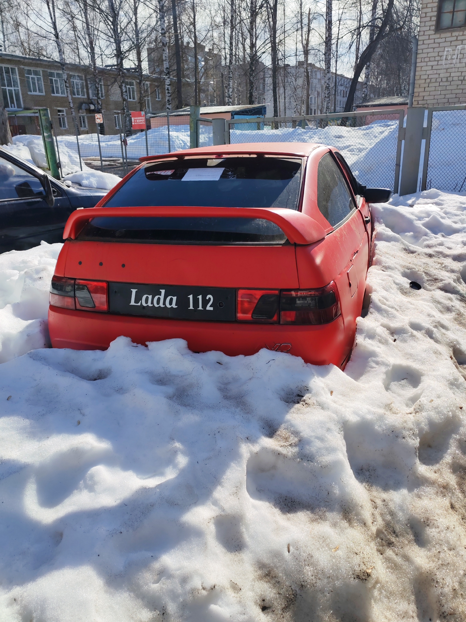 A matte red Lada 112 two-door coupe/hatchback is situated amidst patches of melting, dirty snow under bright sunlight in Chaikovsky, Russia. The vehicle features a rear spoiler, darkened taillights, and "Lada 112" text on a black panel where a standard license plate would be. A section of a dark-colored vehicle is partially visible to the left of the Lada. In the background, a chain-link fence spans horizontally, displaying a red sign with Cyrillic text translating to "DO NOT PARK CARS AT THE GATE" and a red circular no-parking symbol. Behind the fence, a light-colored brick building with numerous windows is visible, partially obscured by numerous bare deciduous trees. To the far right, a segment of a light brown brick wall with hanging icicles is present. Additional multi-story buildings and more bare trees are visible in the distant background. No individuals are present in the scene.