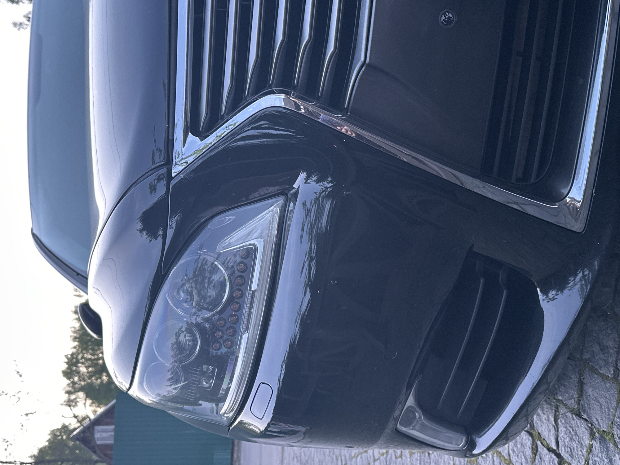The image shows the front passenger side of a dark-colored car, possibly a luxury sedan or SUV, featuring a prominent chrome grille and a modern LED headlight design. The car is likely parked outdoors on a paved surface, potentially cobblestone. In the background, hints of trees and a green building or structure are visible, suggesting a residential or semi-urban setting. The image is taken at ground level, looking upwards towards the front of the car. Новоглаголево, Russia is mentioned, which could be the location where the image was taken. No people are visible in the image.