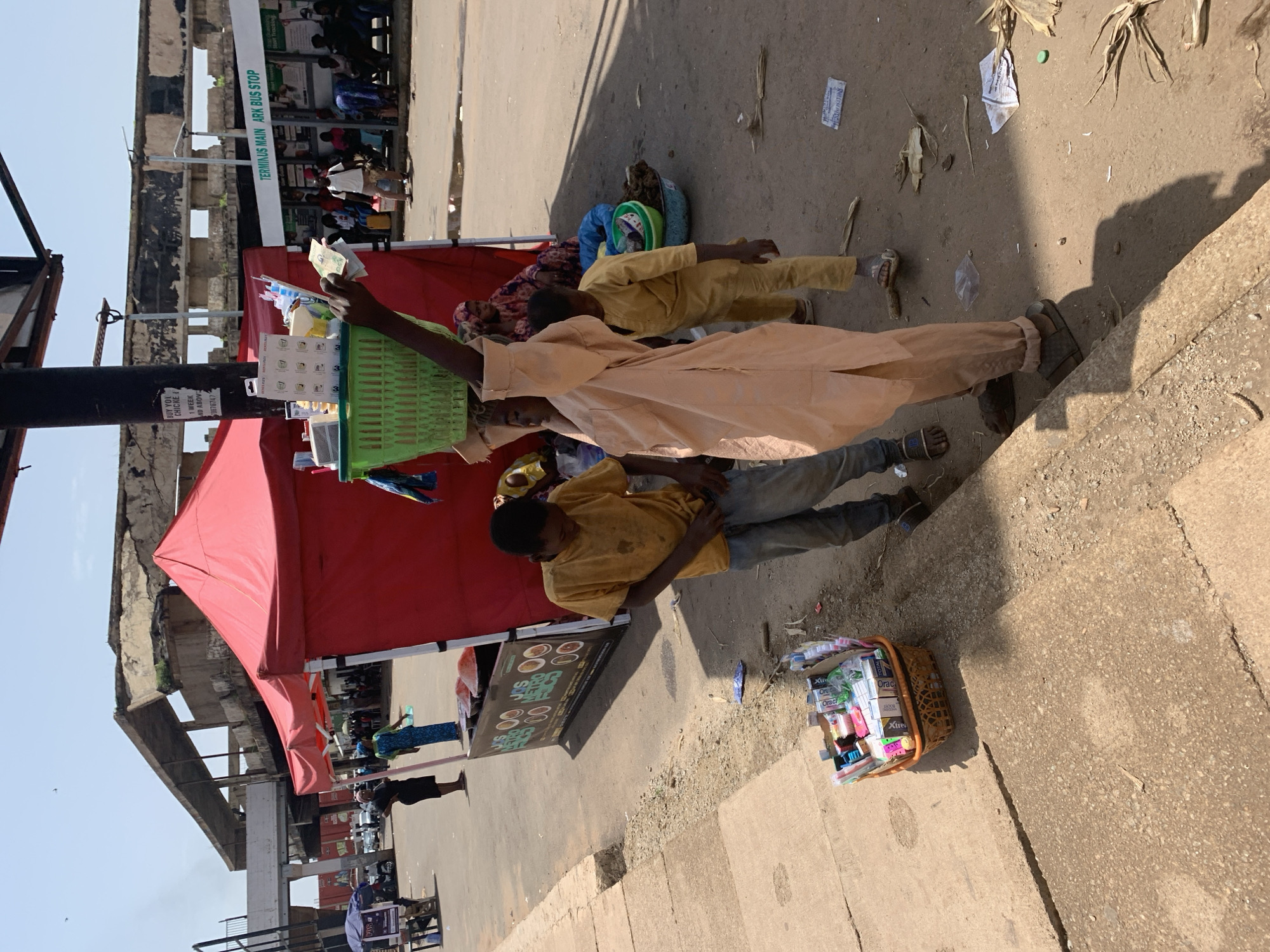The image depicts a street scene in Jos, Nigeria, likely near a transportation hub given the presence of the "TERMINUS MAIN ARK BUS STOP" sign. In the foreground, a young boy in a yellow t-shirt and jeans stands looking down, possibly adjusting his clothing. Next to him, a basket overflowing with toothbrushes and toothpaste is placed on the ground. A taller individual, wearing a light-colored robe, carries a green basket filled with various items, including bottled water and other products, balanced on their head. Behind them, a red tent serves as a makeshift stall. Several other people are visible in the background, some seated, others standing, near the bus stop. The environment appears to be a dusty, open-air market or street, with evidence of dried vegetation and scattered debris on the ground. The overhead structure suggests an elevated roadway or an old, possibly abandoned building.