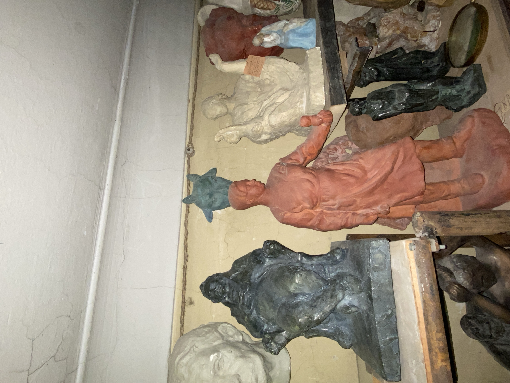 The image depicts a collection of sculptures displayed on shelves against a wall in a dimly lit room. The wall and ceiling show signs of cracking and age. In the foreground, a terracotta-colored statue of a person in what appears to be work clothes stands prominently. To the left, a large, dark-colored bust of a person is partially visible. Behind these, various other sculptures are arranged, including a light-colored figure seated with arms raised, a small blue figure, and a dark green bust with animal-like features. The shelves themselves appear to be made of wood and metal, showing signs of rust and wear. The overall impression is one of a storage or display area for artistic creations. The specific location in Moscow, Russia, cannot be determined from the provided image.