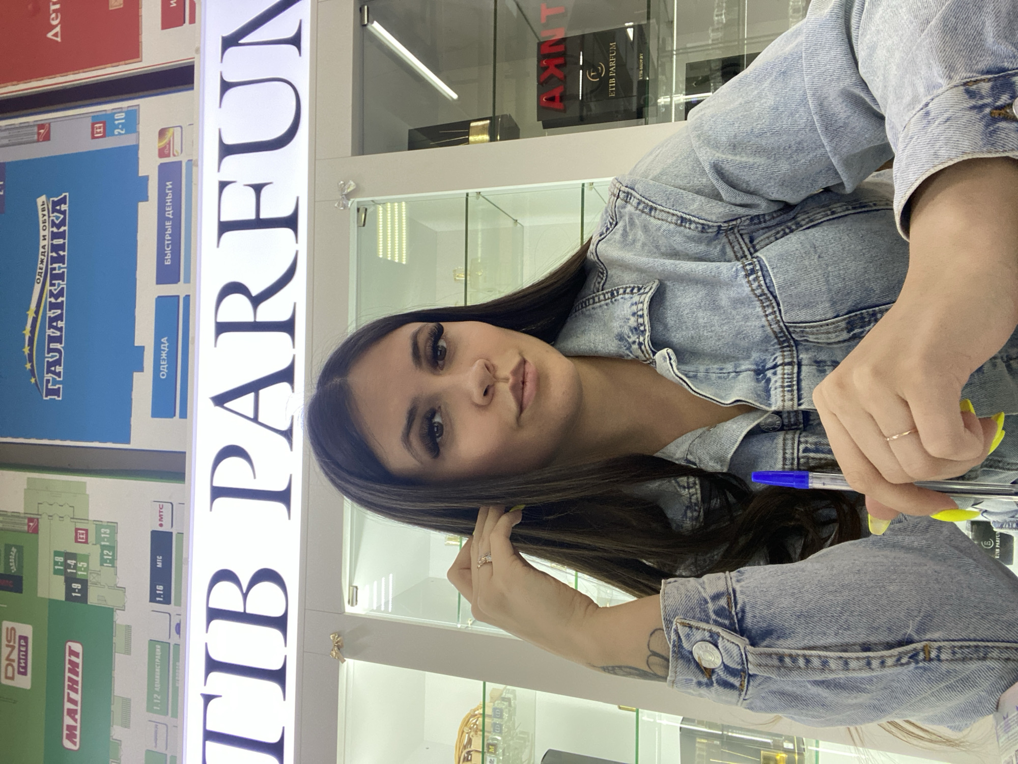 The image depicts a woman wearing a denim jacket, standing in front of a storefront sign that reads "TIB PARFUM". Adjacent to the sign is a directory board with various labeled sections and store names, including "DNS ГИПЕР", "МАГНИТ", and "ГАЛАКТИКА". Based on the Russian text "Чайковский", this scene is located in the city of Chaikovsky, Russia. The woman is holding a pen in her hand and appears to be in a retail environment, as indicated by the perfume brands "ETIB PARFUM" visible behind her in the glass display cases.