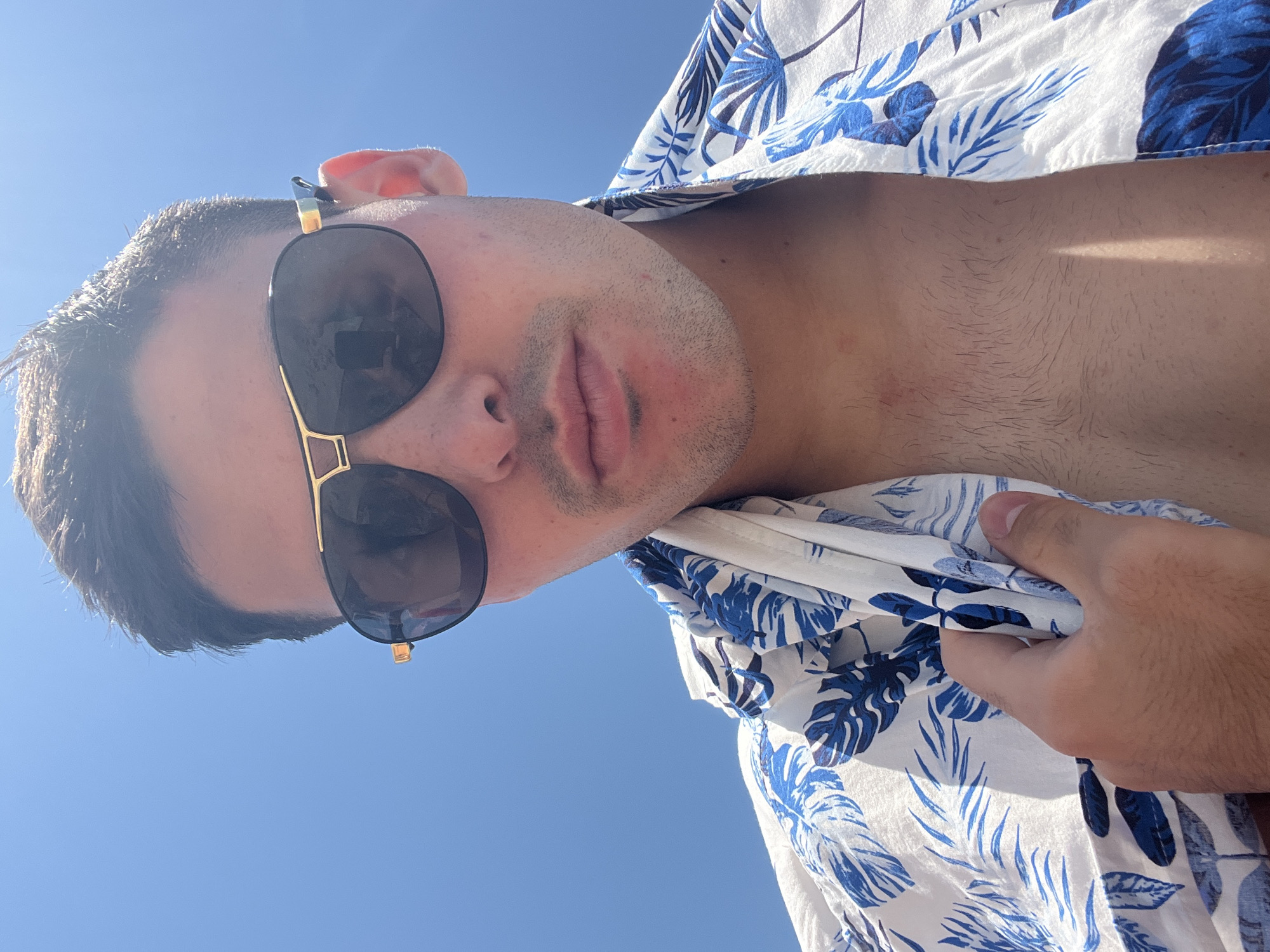 A low-angle, rotated portrait captures a person from the chest up against a clear, bright blue sky. The individual, appearing to be a young adult male, has short, dark hair and light stubble on his face. He wears dark aviator-style sunglasses with gold-colored frames, which reflect the intense sunlight. His lips are slightly parted, conveying a relaxed expression. He is dressed in a short-sleeved, open-collared shirt featuring a white background adorned with a blue tropical leaf pattern (resembling palm or fern fronds). A hand is visible on the right side of the frame, gripping the fabric of his shirt. The strong, direct sunlight and lack of clouds indicate a clear, sunny daytime, likely mid-day, in an outdoor setting. The perspective looks upward towards the subject, with the expansive blue sky dominating the background. No discernible text is visible.