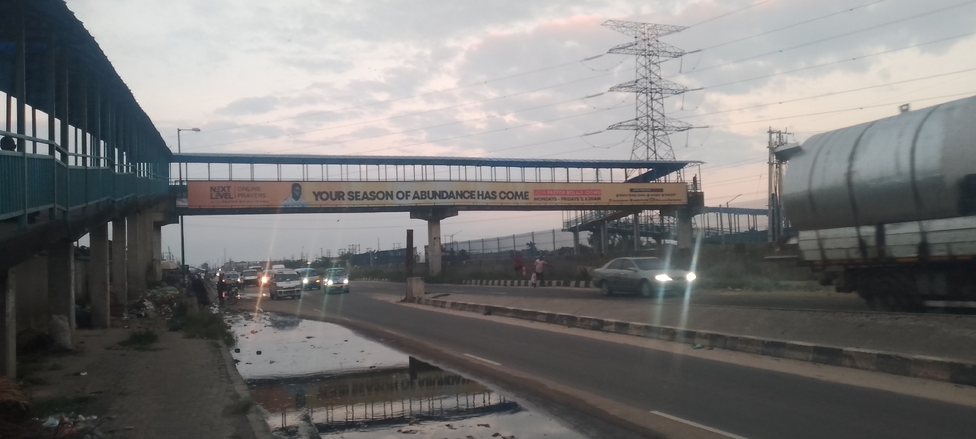 The image depicts an overcast sky with scattered clouds, suggesting either dawn or dusk. A pedestrian bridge, with a blue railing and a silver roof, spans over a multi-lane road. A large banner hangs from the bridge, featuring text and a portrait of a man. The banner reads "NEXT LEVEL PRAYERS" and "YOUR SEASON OF ABUNDANCE HAS COME JOIN PASTOR BOLAJI IDOWU MONDAYS - FRIDAYS 6:30AM". Traffic is present on the road, including cars and a large tanker truck. The roadside is lined with debris and a puddle of water reflecting the overhead structures and sky. In the background, a large electrical pylon and industrial-looking structures are visible. A few individuals are standing on the side of the road near the pedestrian bridge. The overall scene appears to be an urban or semi-urban environment.