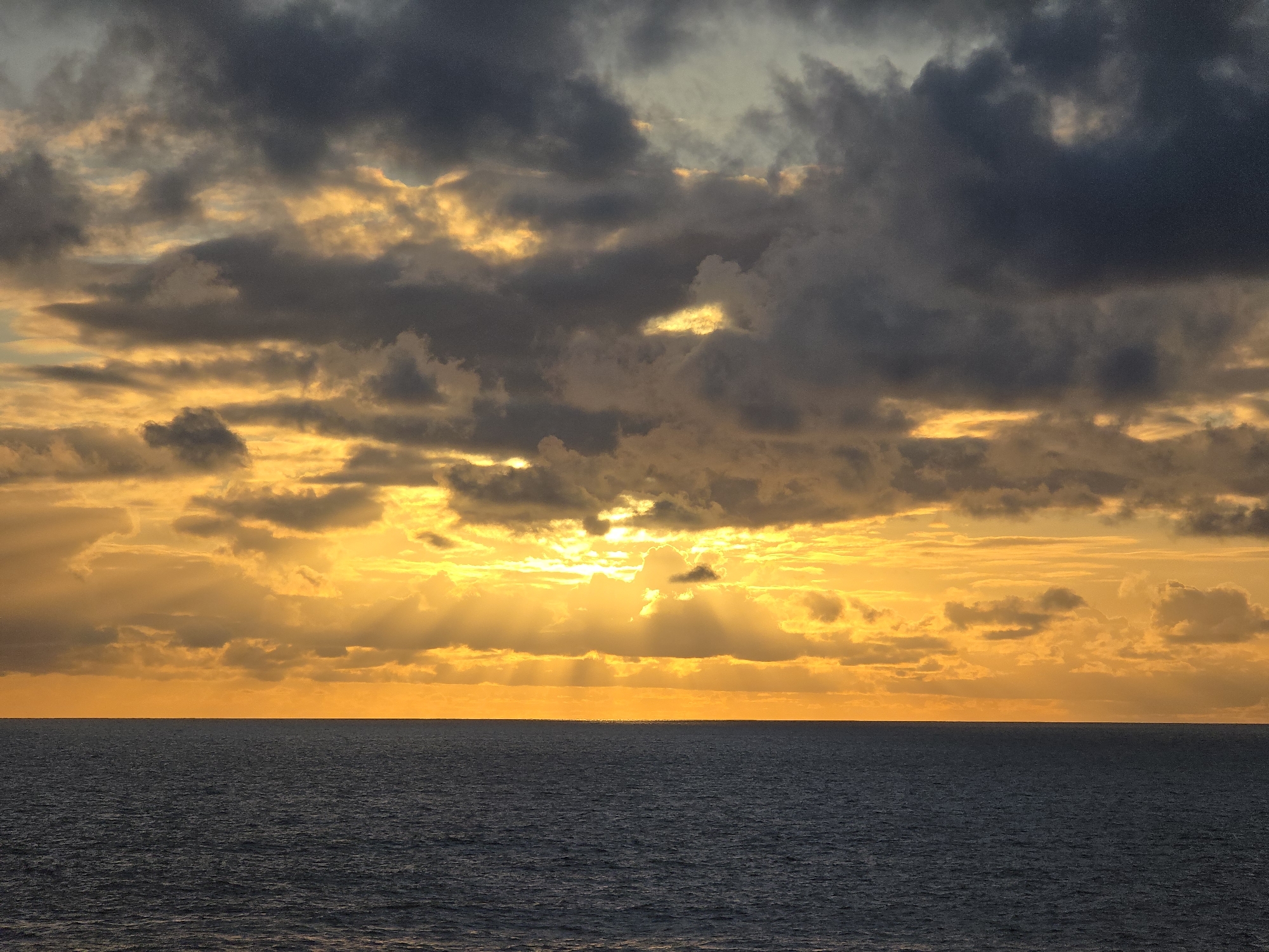 The image depicts a sunset over the ocean. The sky is filled with dark, heavy clouds illuminated by the setting sun, which creates a vibrant orange and yellow glow. Sunbeams pierce through the gaps in the clouds, casting light onto the water below. The horizon line is clearly visible, separating the sky and the sea, which appears dark and calm. There are no people or specific objects identifiable within the frame. The location is the ocean in Unknown, Unknown.