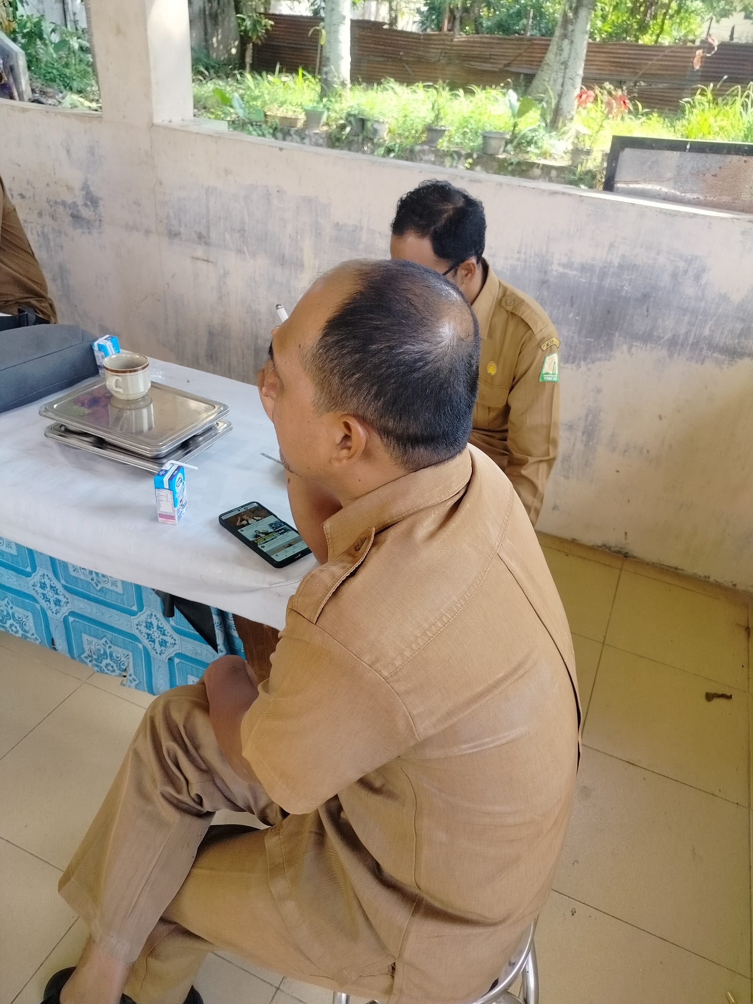 In Purwodadi, Indonesia, two men in tan government uniforms are seated at a table covered with a white tablecloth. The man in the foreground, seen from behind and the side, is bald and appears to be smoking. He is looking down at a smartphone on the table, which displays content that is not clearly discernible. Beside the phone is a small carton of milk with a straw. A mug sits on a metal tray on the table. The second man, in the background, is partially visible, also wearing a tan uniform with a patch on his sleeve. The setting appears to be an outdoor covered area, with a concrete wall behind the table and vegetation visible in the distance. The floor is tiled.