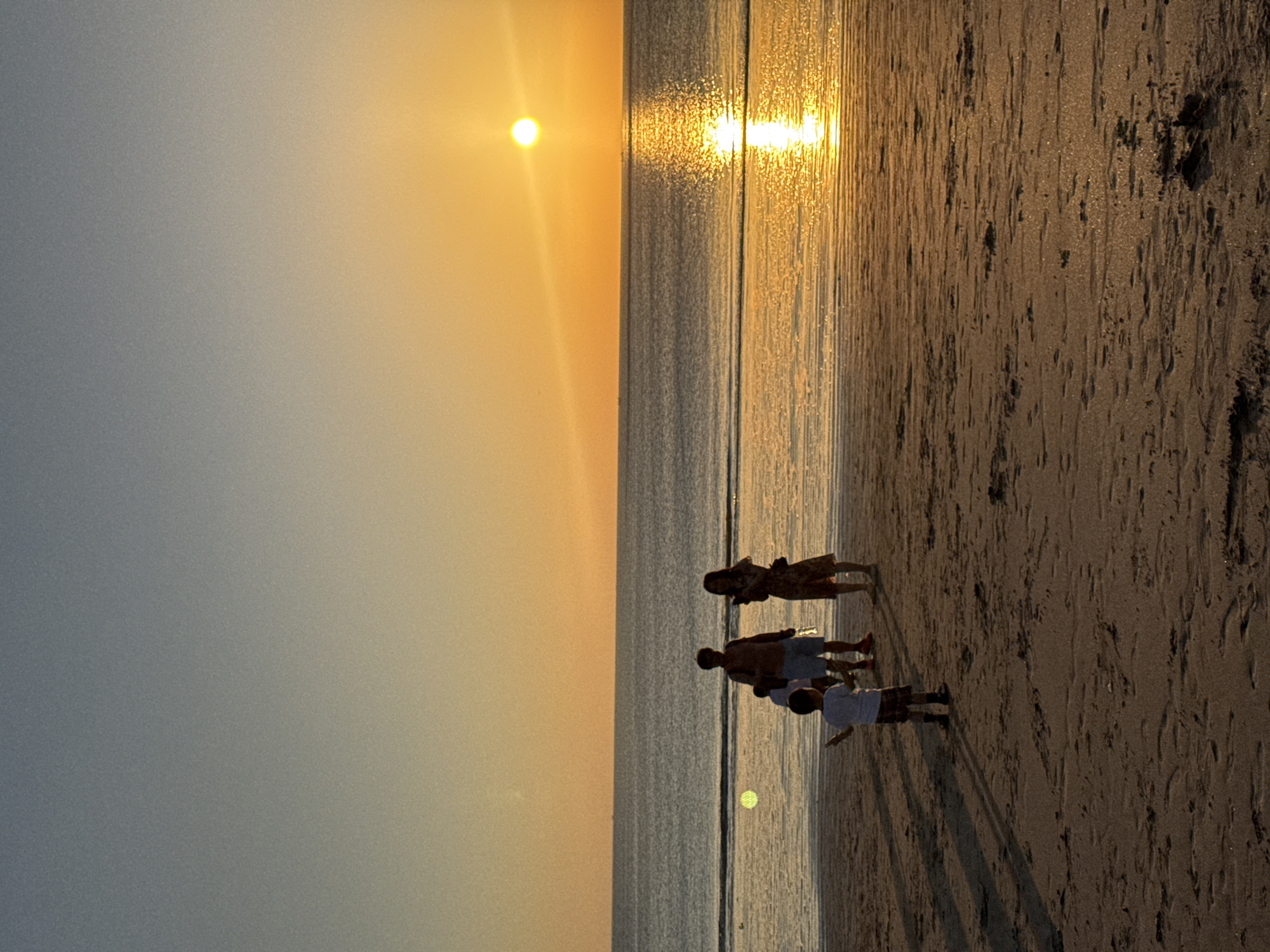 A family of four is silhouetted against a sunset on a sandy beach. The sun is low on the horizon, casting a golden glow across the sky and water. The ocean is calm, with gentle waves lapping at the shore. The sand is wet and dark near the water's edge, transitioning to a lighter, drier tone further up the beach.
The family members are standing close together, facing the water. A woman in a patterned dress is on the left, followed by a man in shorts and a t-shirt. Two younger figures, likely children, are to the right of the man. Their stances suggest they are enjoying the view or perhaps interacting with each other. Long shadows stretch from the family onto the sand, indicating it is late afternoon or early evening.
The scene evokes a sense of peace and togetherness. The warm colors of the sunset and the tranquil atmosphere suggest a relaxing vacation. The location is identified as Kuta Selatan, Indonesia, a popular tourist destination known for its beaches and sunsets. There is no visible text in the image.