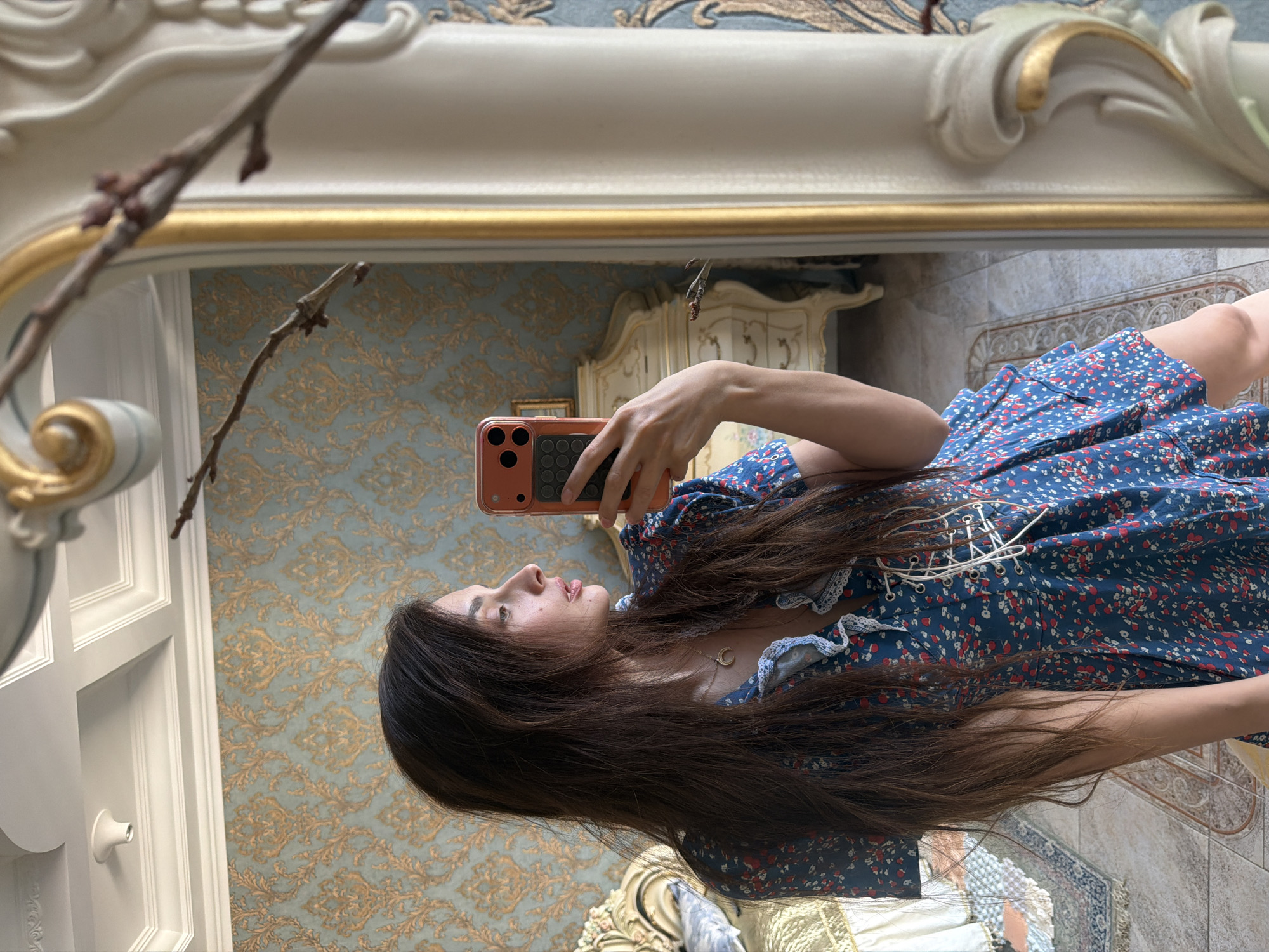 The image shows a young woman taking a mirror selfie in a room with ornate wallpaper. The woman has long, dark brown hair and is wearing a blue dress with a red and white floral pattern and lace trim. She is holding up an orange cell phone to take the picture. In the background, a gilded mirror reflects the room, which appears to be a bedroom with a dresser and a large wardrobe. The wallpaper is a pale blue with a gold damask pattern. A branch with small buds is visible in the foreground, leaning into the frame. The overall impression is one of a personal moment captured in a luxurious and possibly vintage setting. The lighting suggests it might be daytime, though there are no direct windows visible. The woman's expression is neutral. There is no visible text. The location context provided is Ufa, Russia.